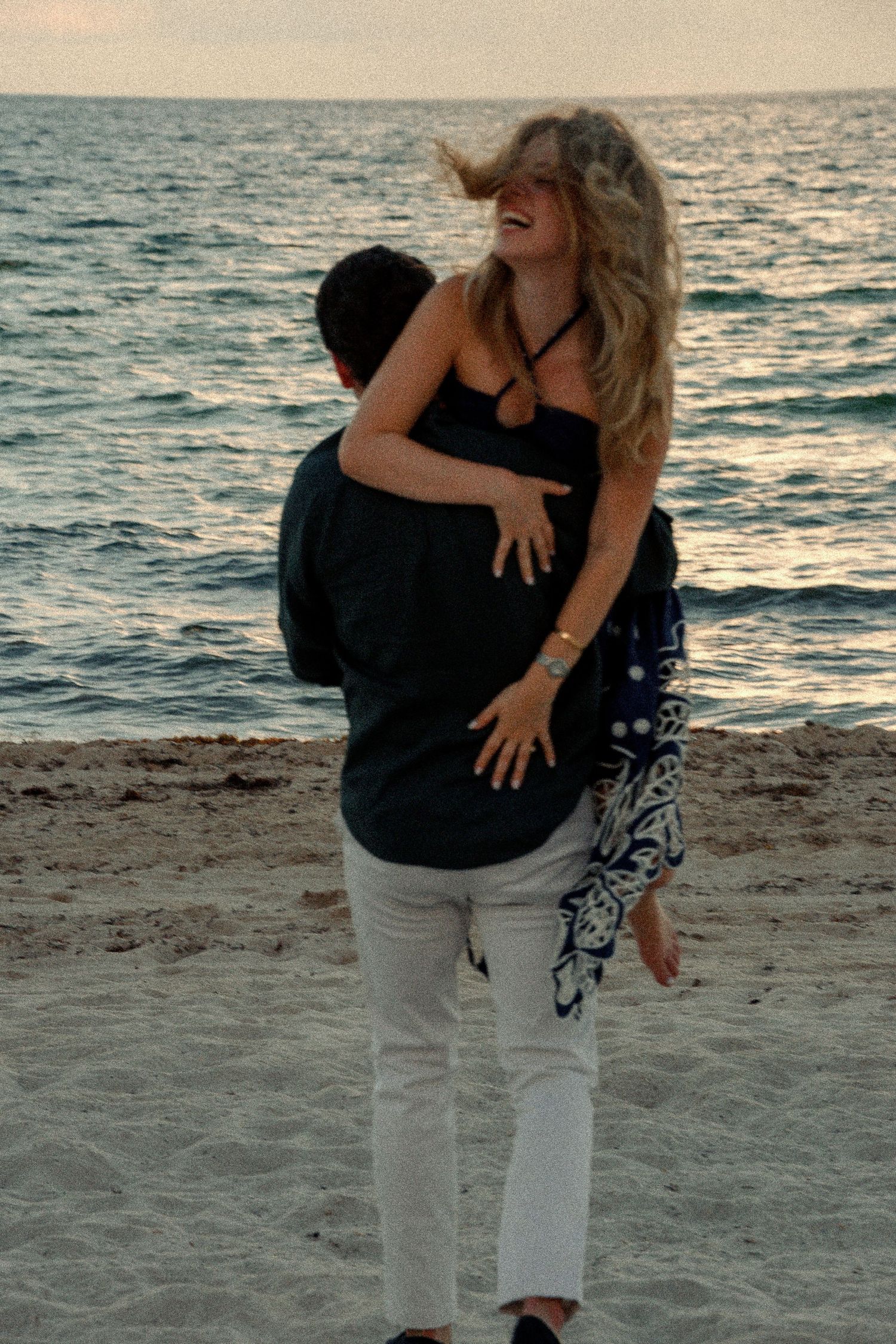 Couple embracing on a sandy beach at sunset, woman with blonde hair laughing as man lifts her up.