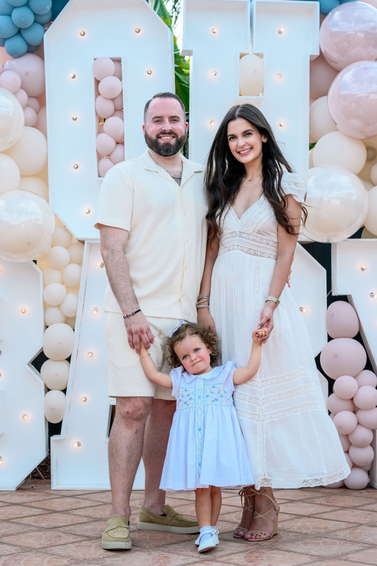 A family of three poses in front of a balloon arch display, parents holding hands with their toddler daughter in a blue dress.