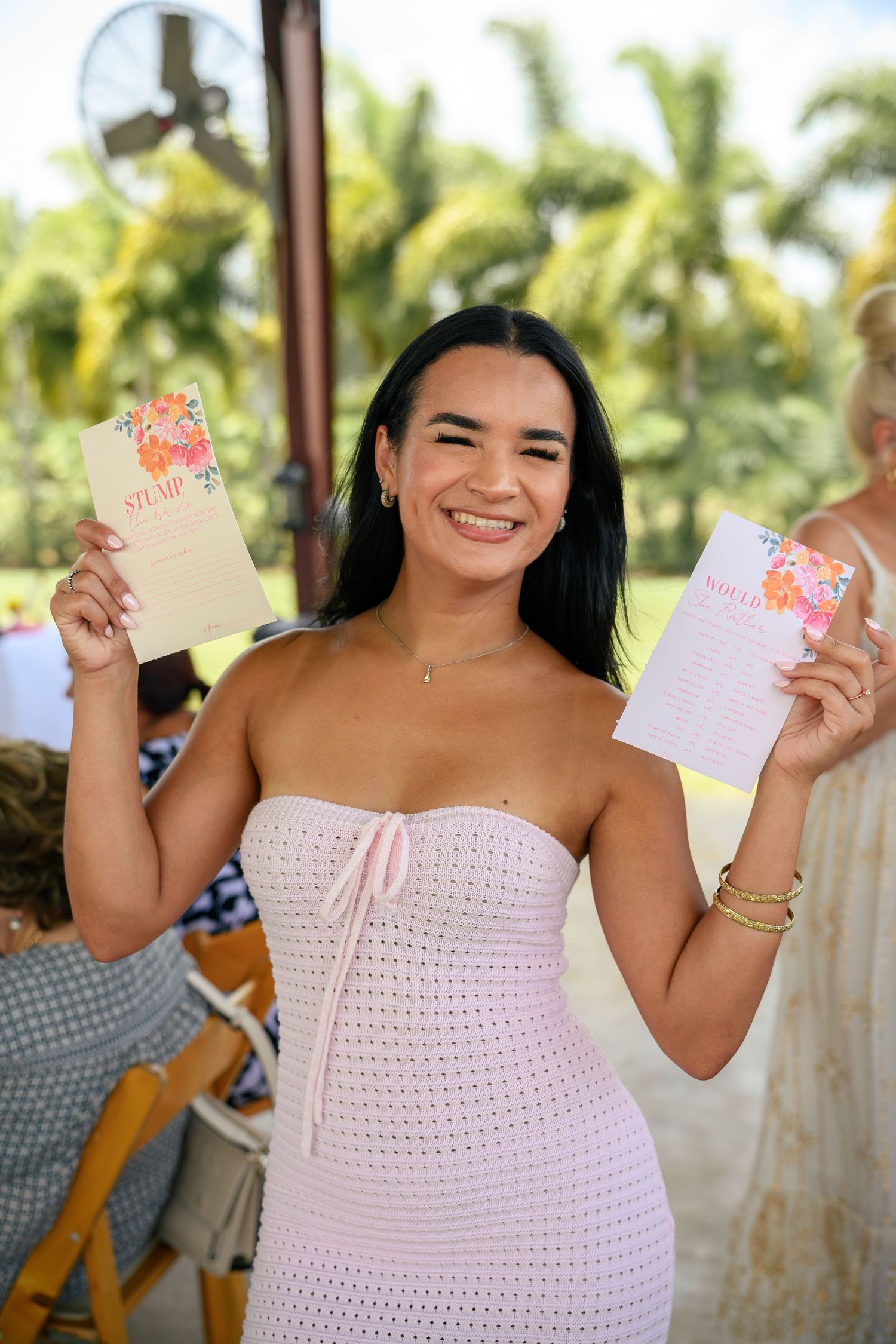 Someone in a white strapless dress holds up two greeting cards at an outdoor tropical venue.