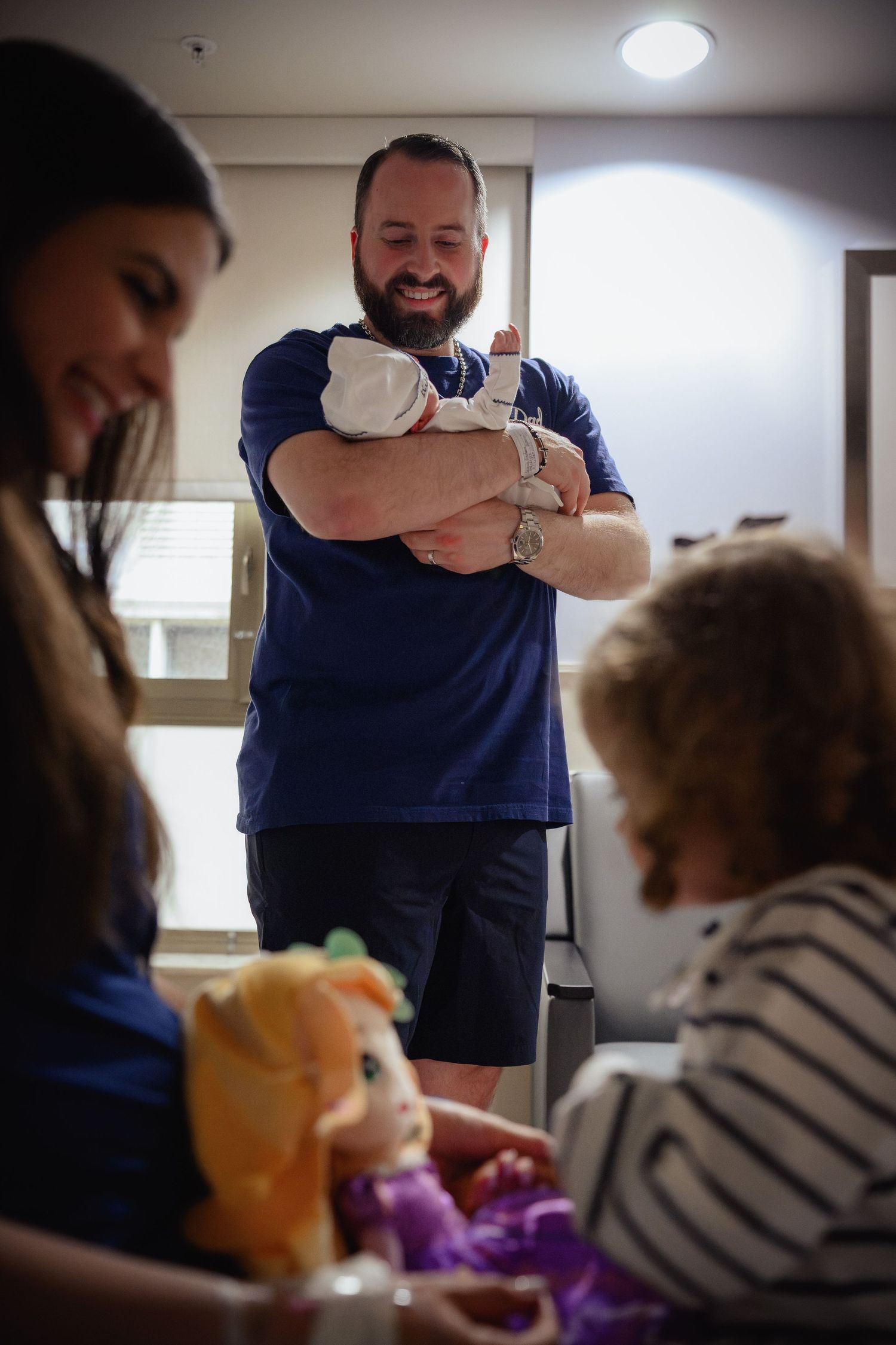 A bearded man proudly holds a newborn baby in a hospital room while family looks on.
