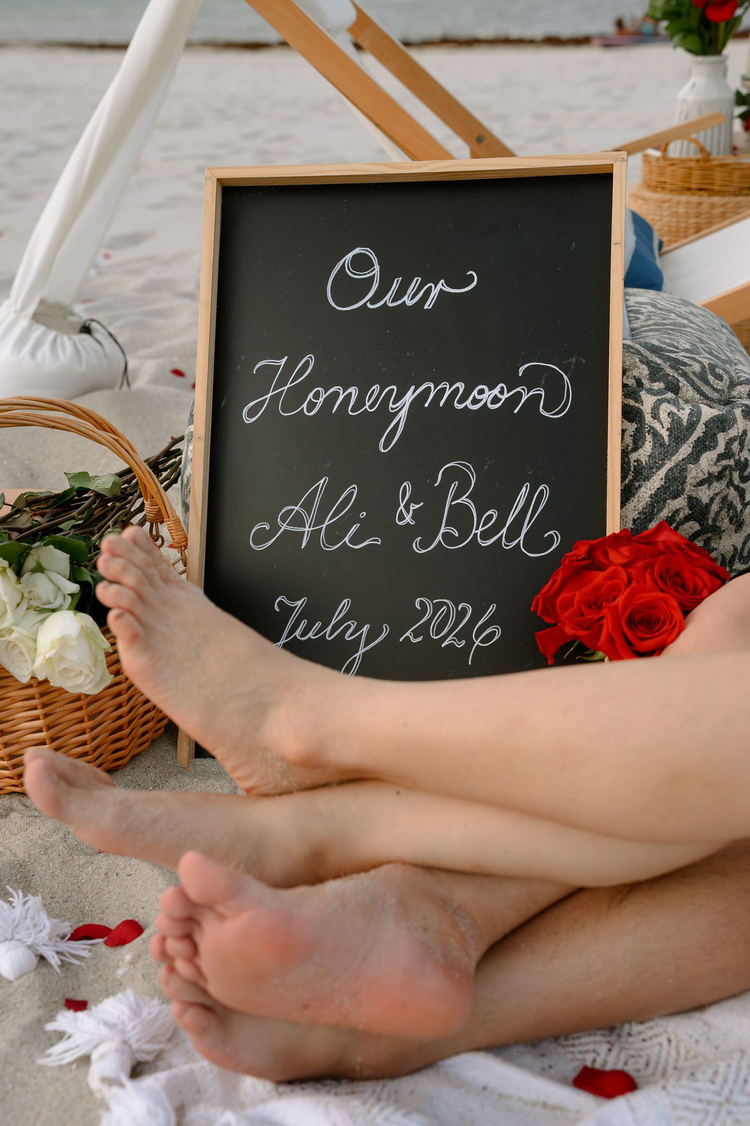 Honeymoon chalkboard sign for Ali and Bell, July 2026, on a beach blanket with roses and a picnic basket.