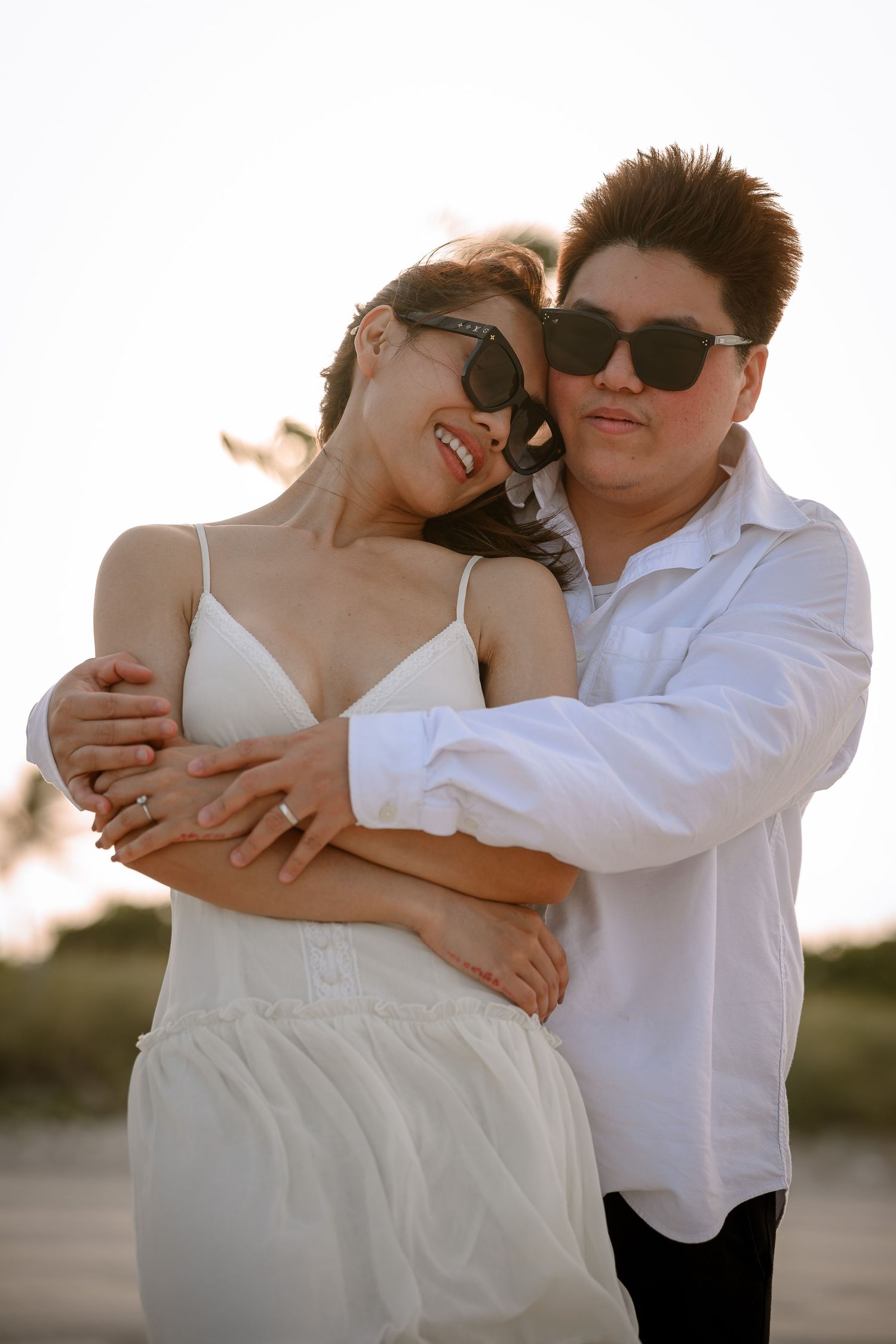 Couple in sunglasses embracing on beach, woman in white dress leaning back against partner in white shirt at sunset.