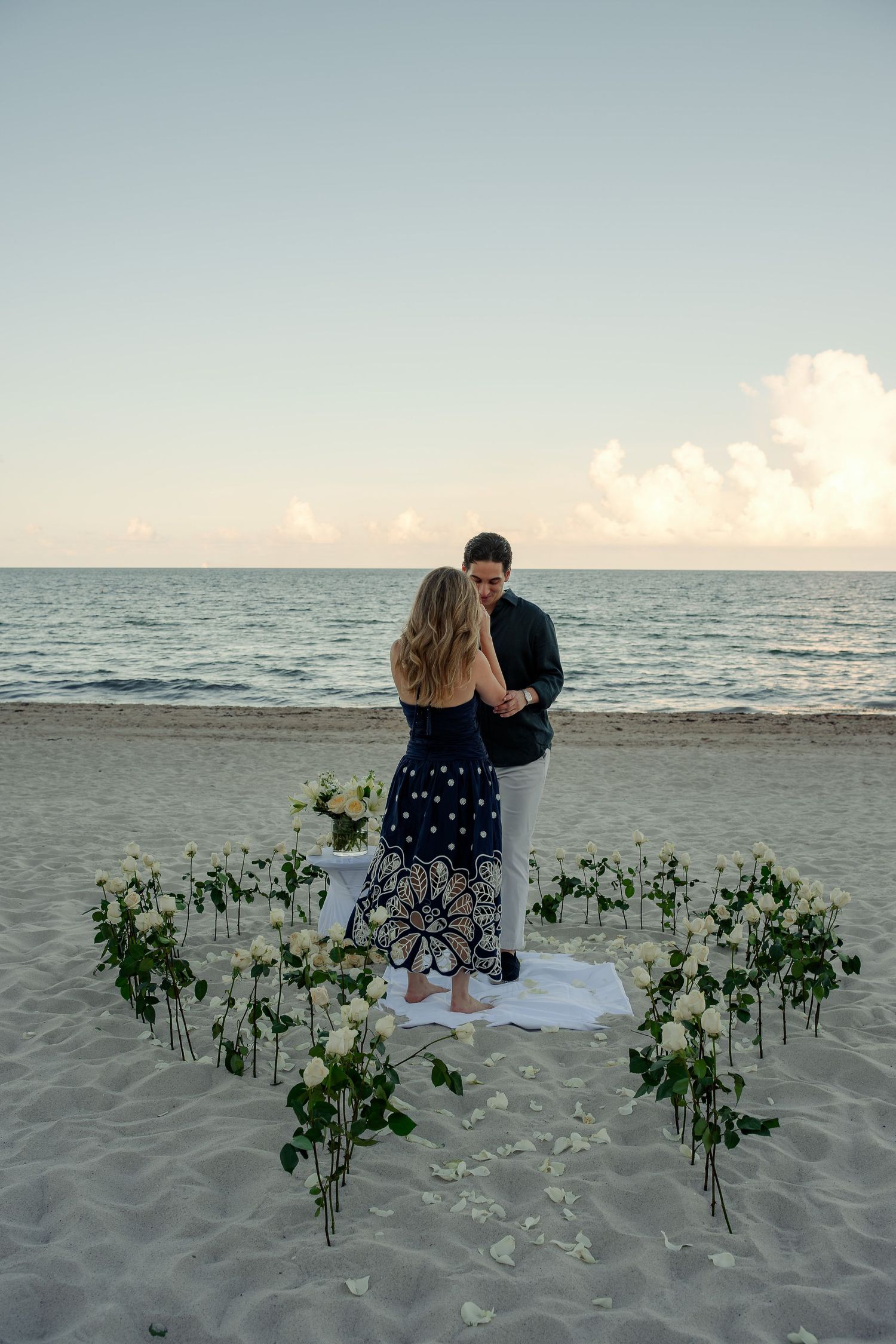 Couple embracing on a beach surrounded by white roses arranged in a heart shape at sunset.