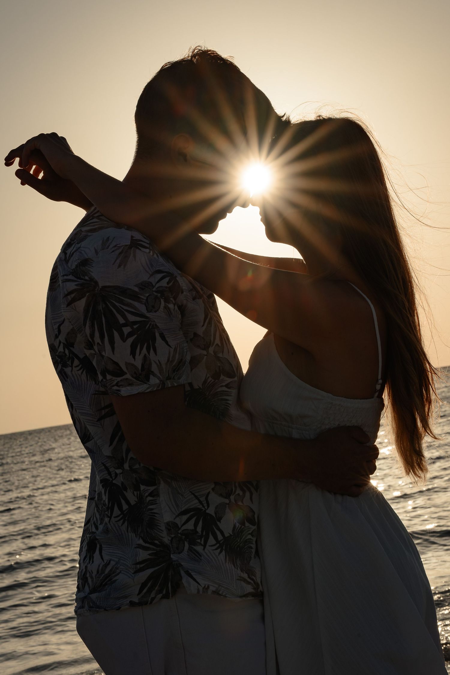Romantic couple silhouetted against a stunning sunset over the ocean, embracing with a sun flare between them.