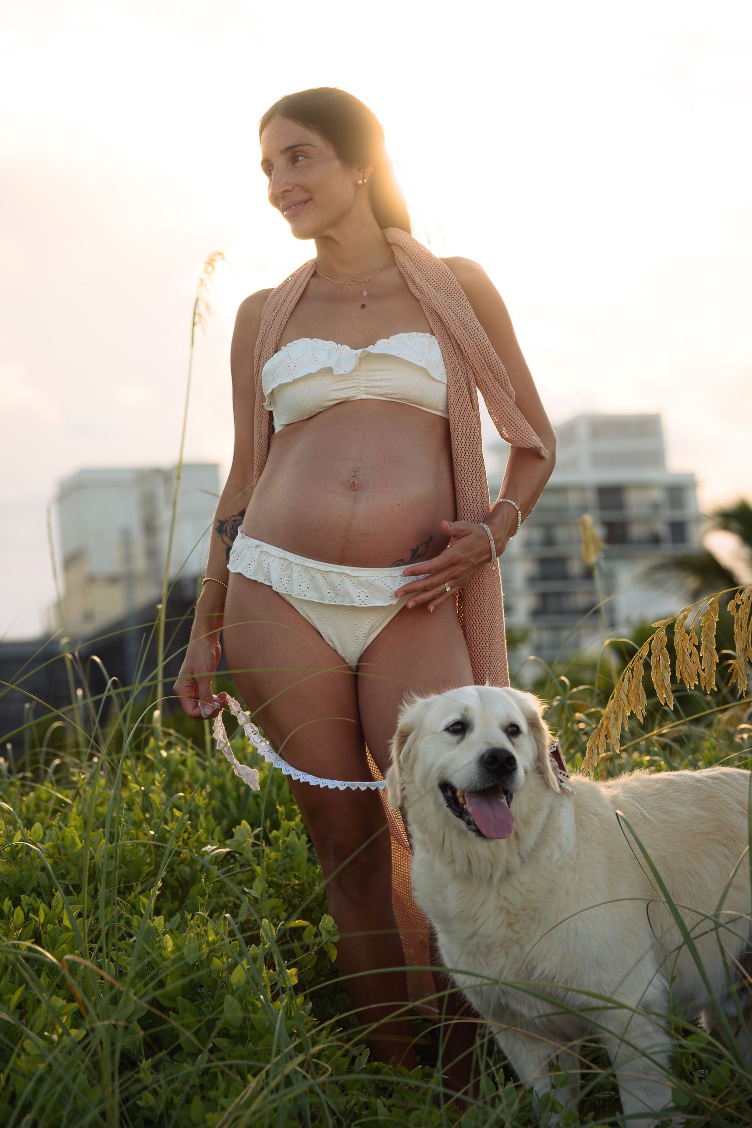 Pregnant woman in white bikini stands in coastal grass with golden retriever at sunset.
