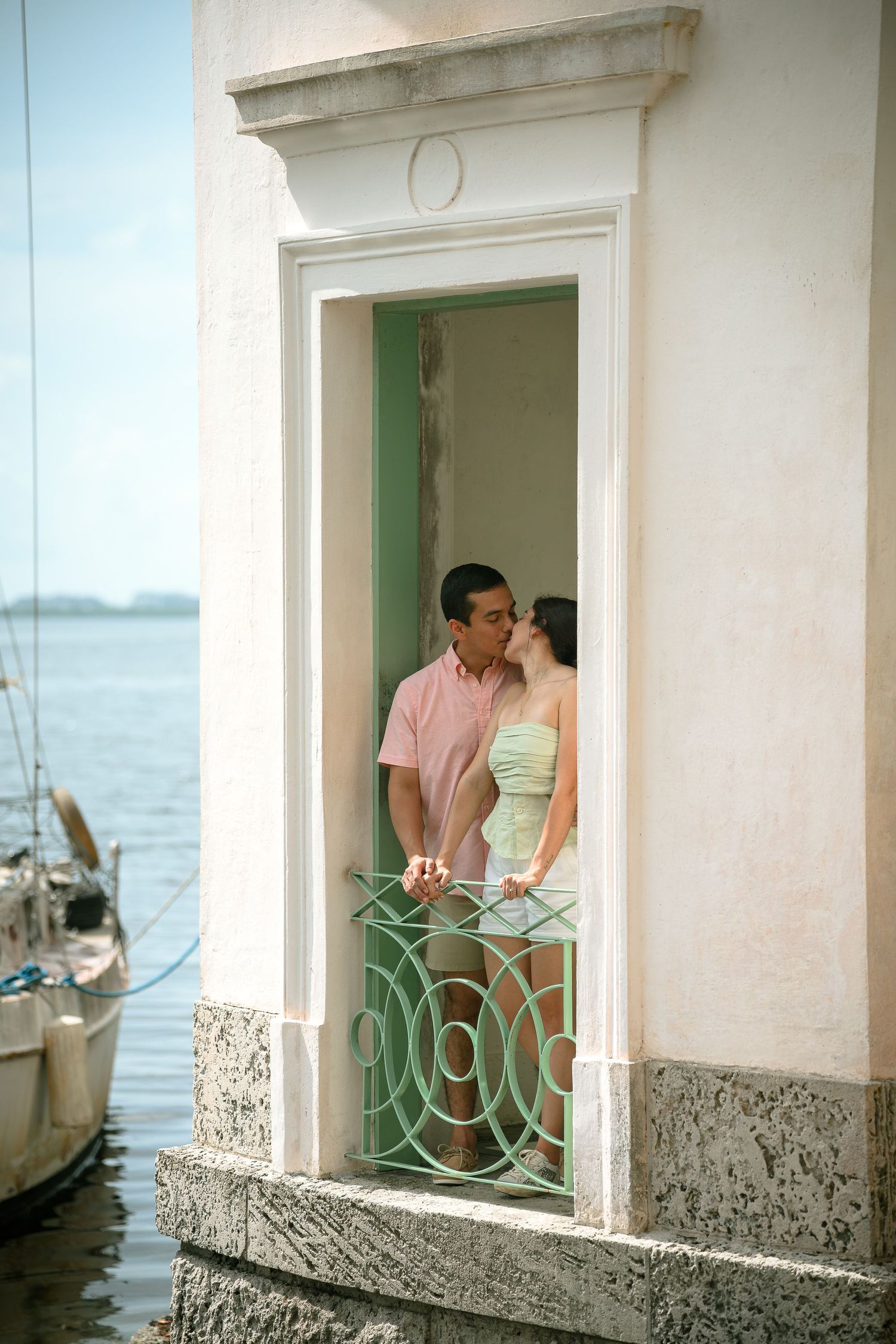 Couple sharing a romantic kiss on a vintage green balcony by the waterfront, framed by elegant white architecture.