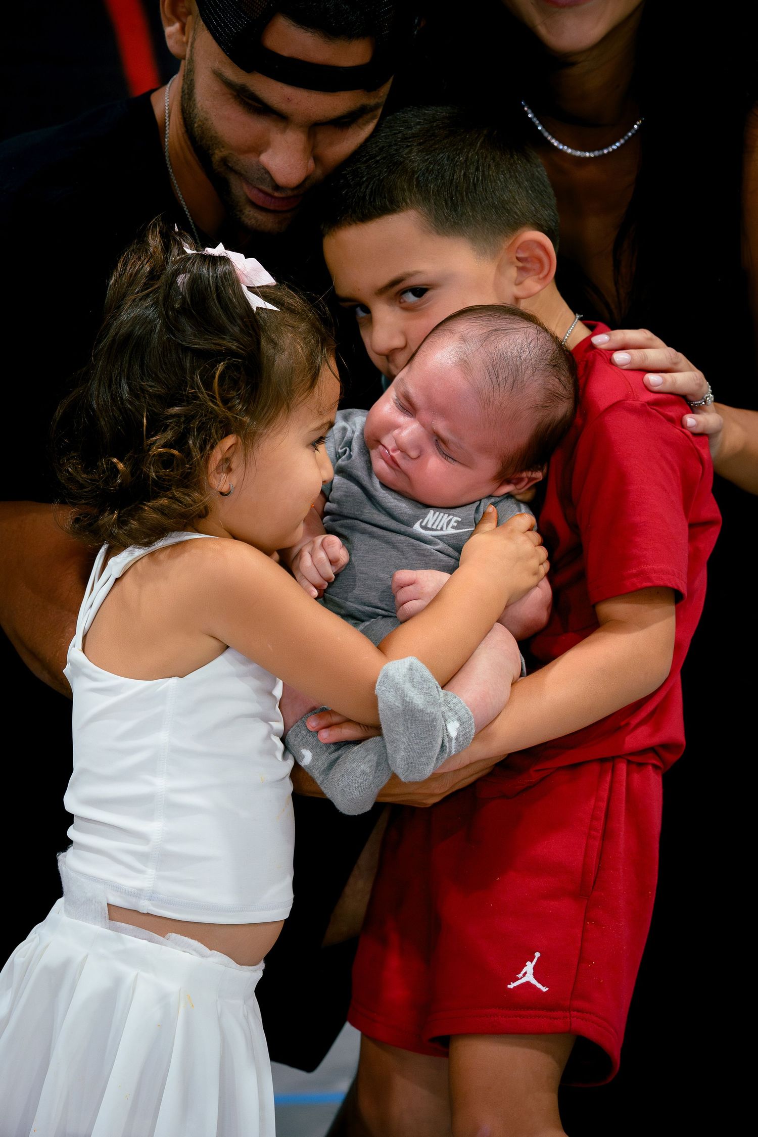 Two children and an adult holding a crying newborn baby dressed in a Nike onesie at a family gathering.