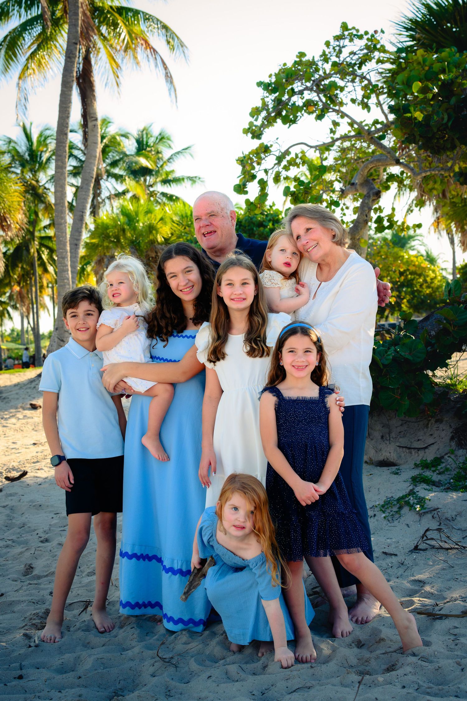 A group in coordinating blue and white outfits poses together on a tropical beach with palm trees at sunset.