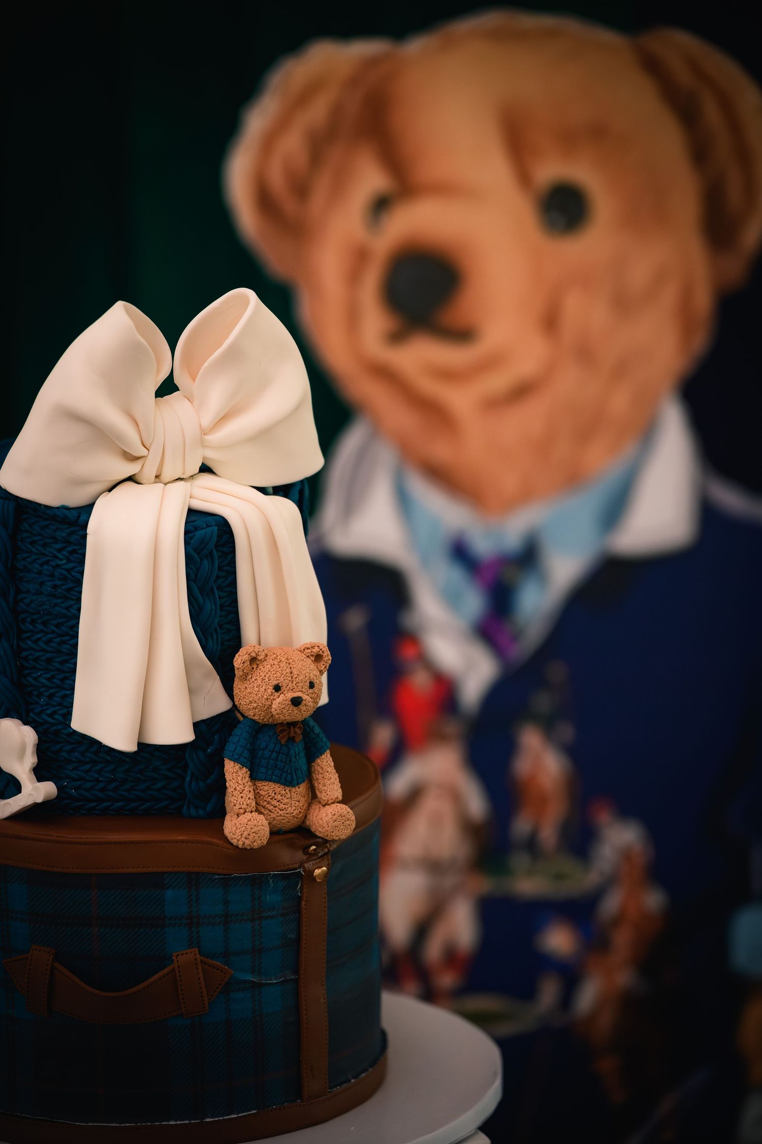 Decorative baby shower cake with tartan pattern, white bow, and small teddy bear topper in front of bear display.