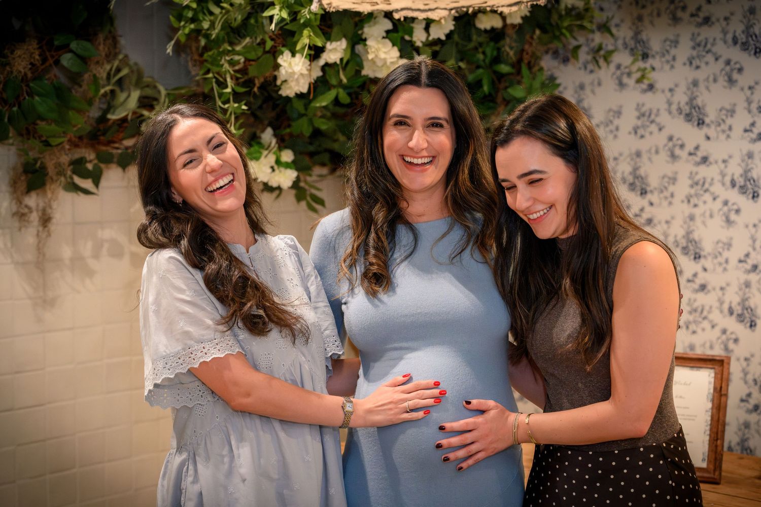 Three friends in light colored dresses share a joyful moment together at a baby shower celebration.