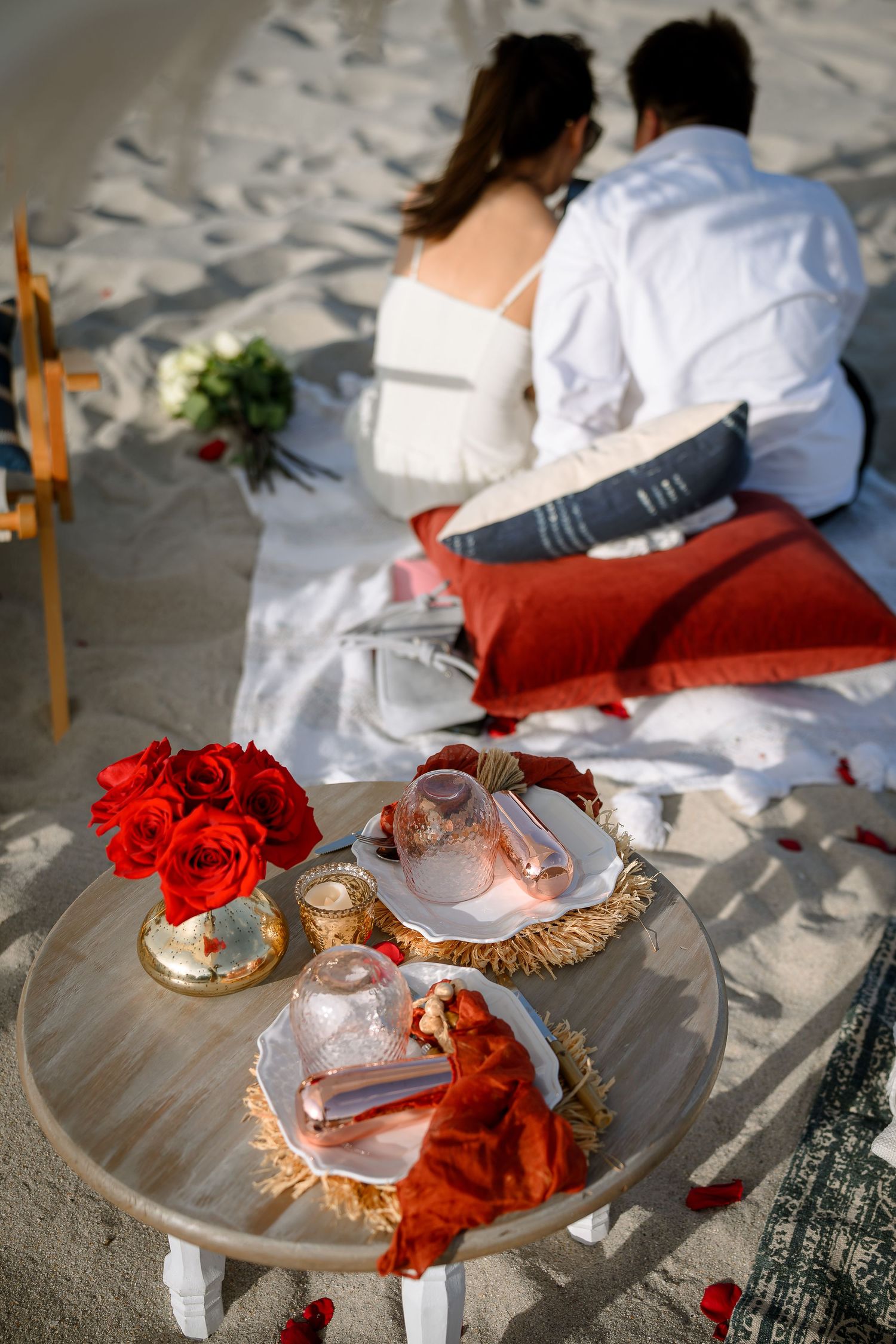 Romantic beach picnic setup with red roses, candles, and a couple sitting on white blanket with red pillows at sunset.