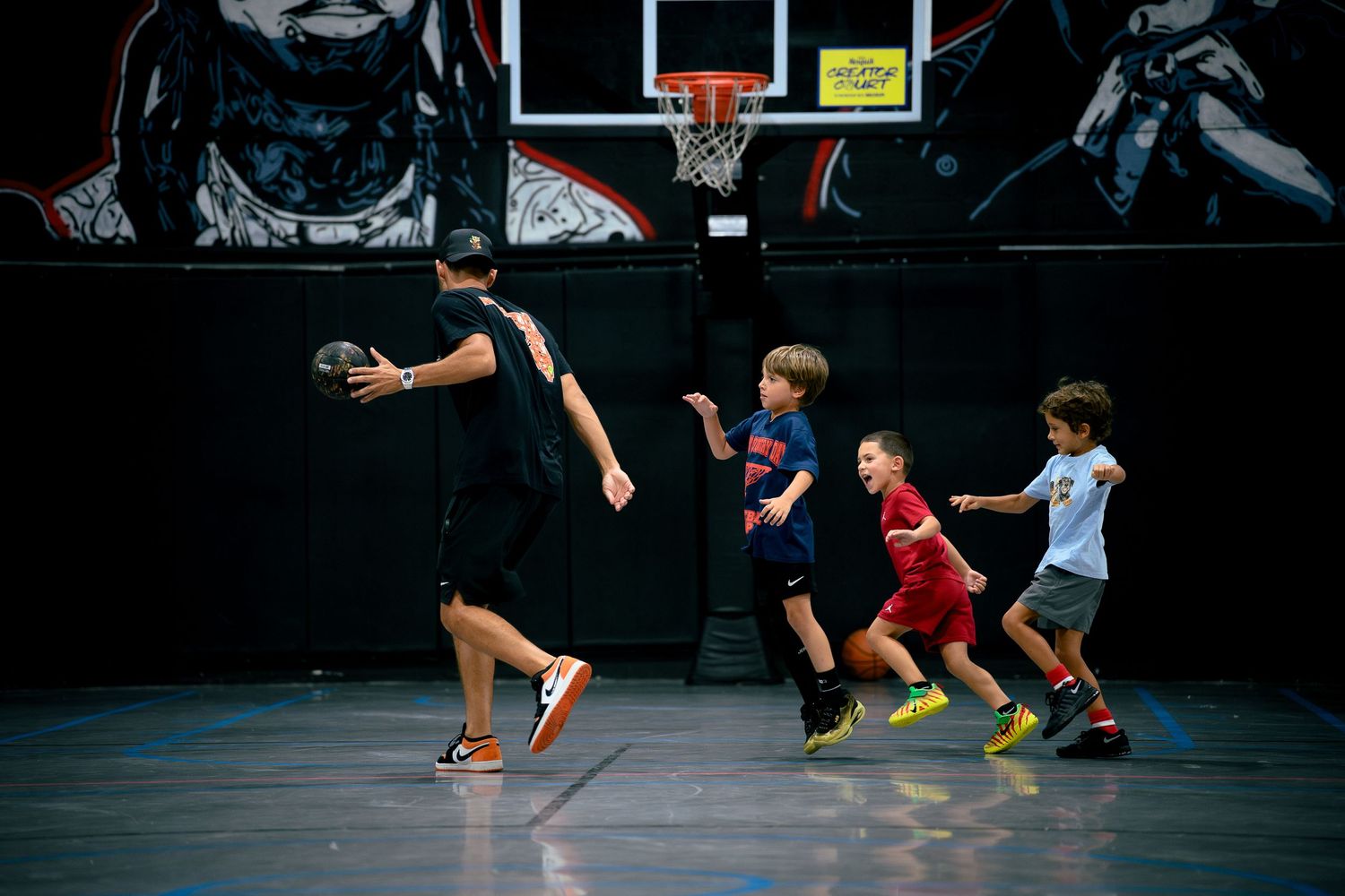 Basketball coach dribbling past three young children during indoor youth basketball training session.