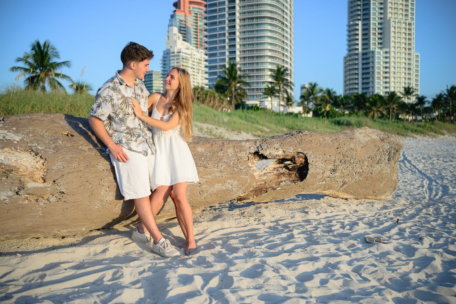 Romantic couple embracing on a sandy Miami Beach with palm trees and luxury high-rise buildings in the background.
