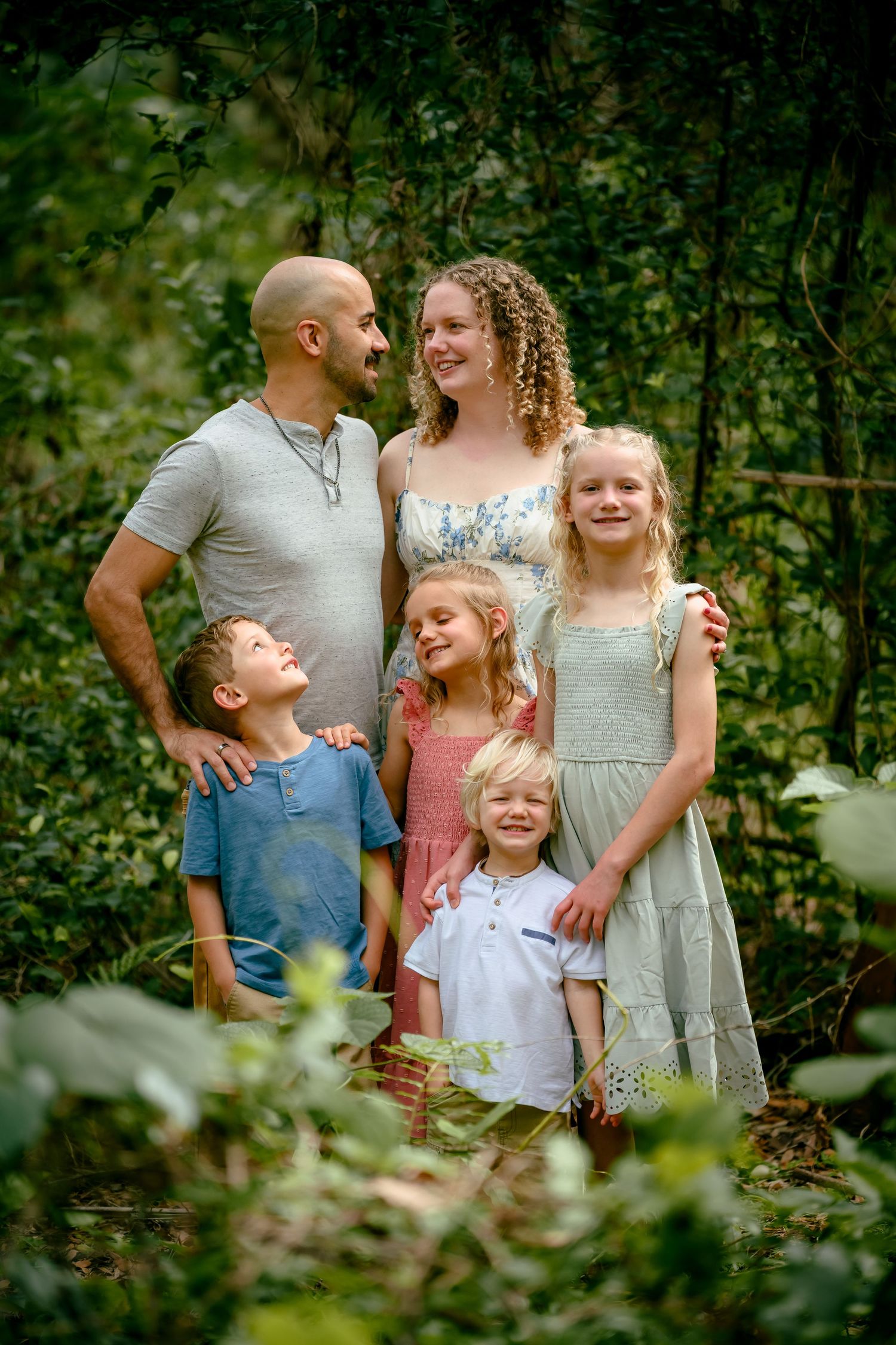 A happy family of five poses together surrounded by lush green foliage outdoors.