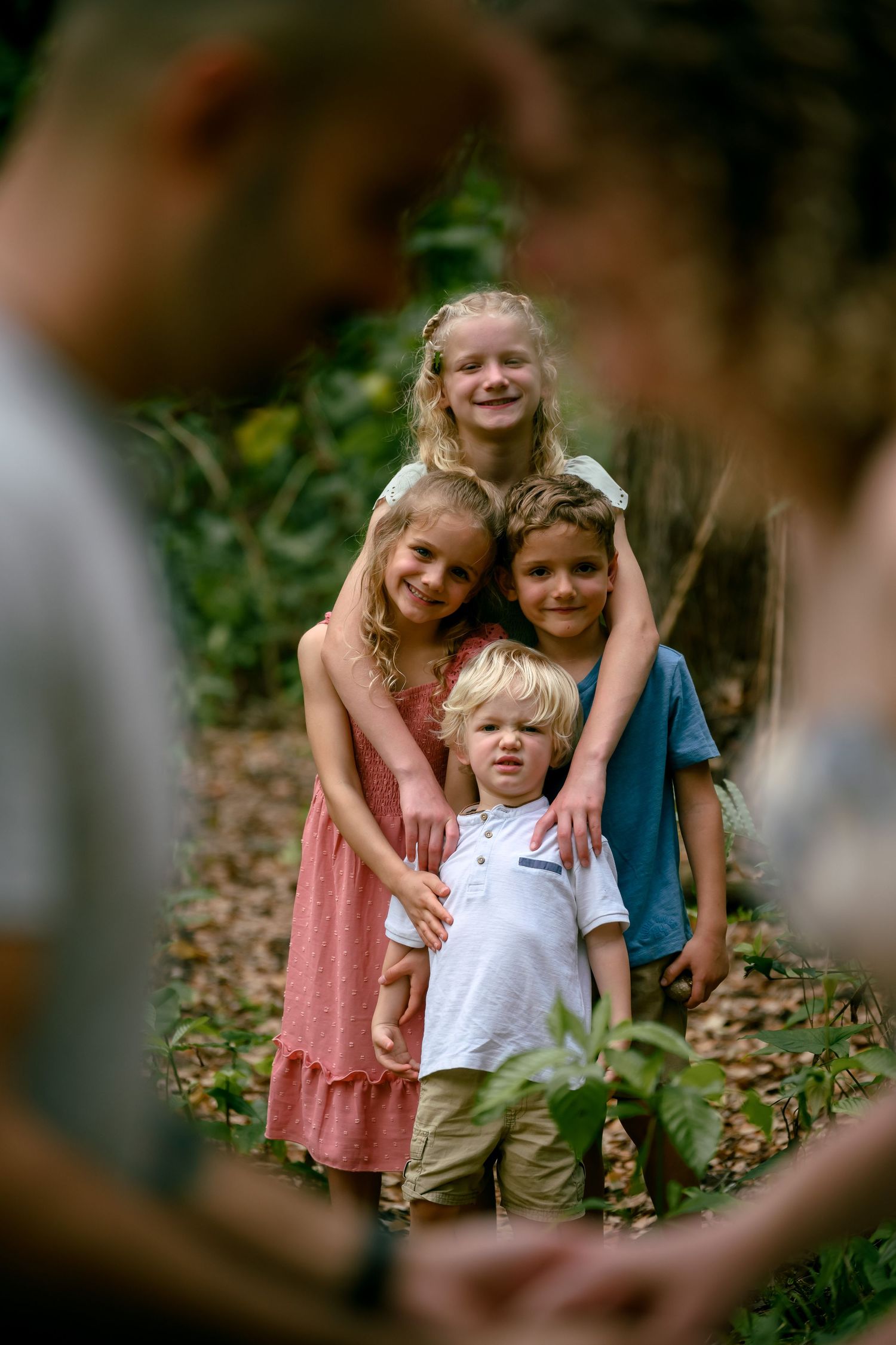 Four children pose together in a wooded outdoor setting, smiling warmly with lush green foliage in the background.