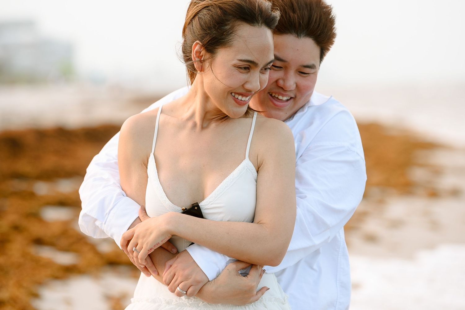 Two women in white embrace and laugh joyfully on a beach, appearing to celebrate a wedding.