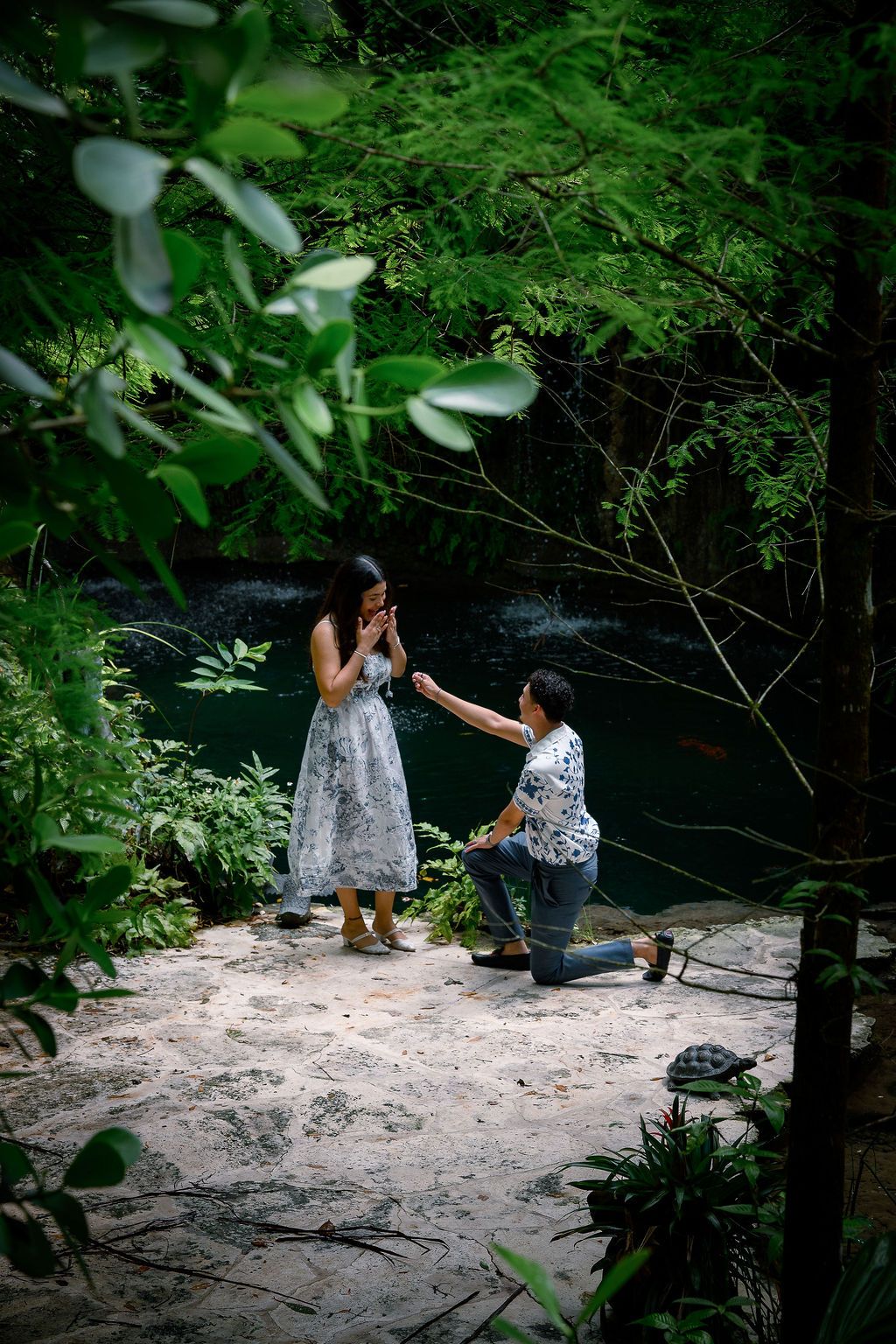 Man proposing to woman on one knee in a lush green forest setting near a river, romantic outdoor marriage proposal.