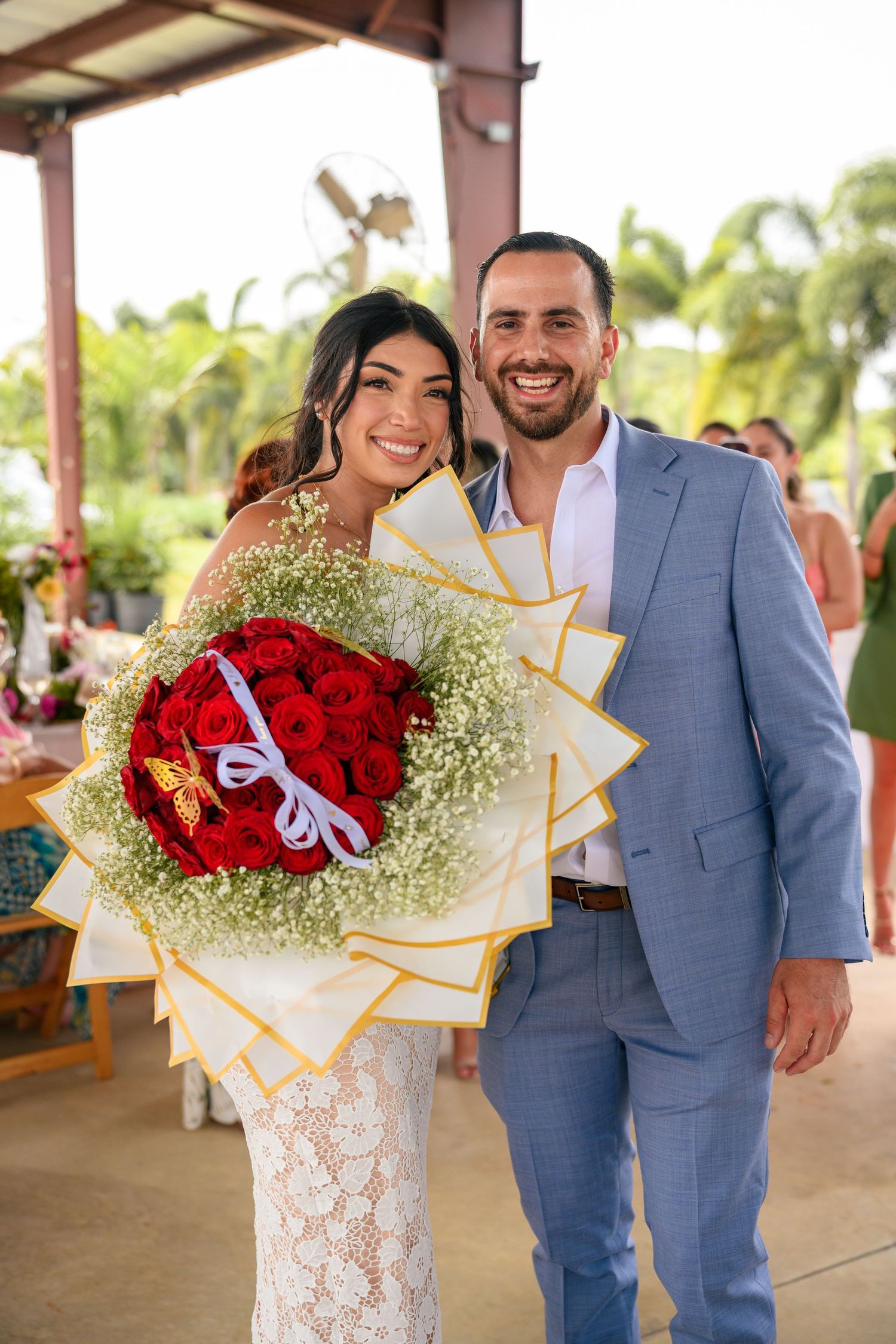 A couple poses outdoors with a vibrant red rose and baby's breath bouquet at a wedding venue with palm trees.