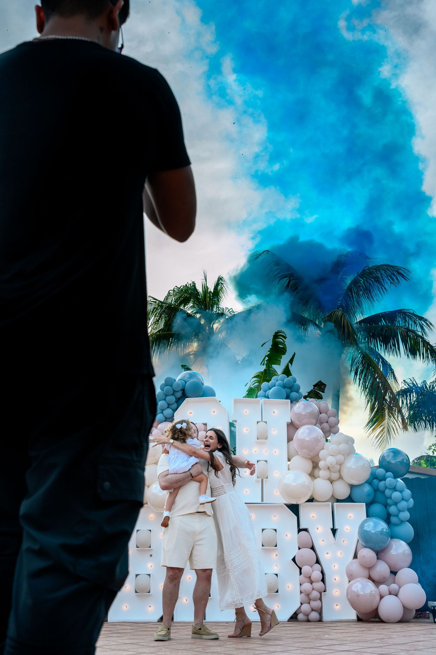 White balloon arch and tropical palm trees create an elegant outdoor wedding ceremony setup against a bright blue sky.