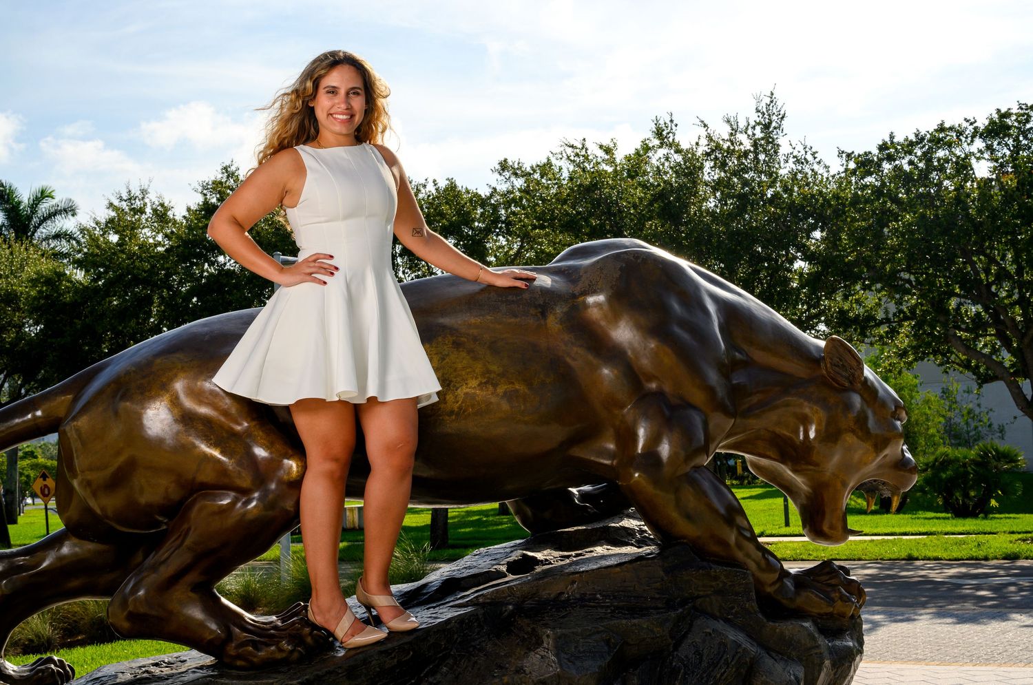 Young woman in white dress posing confidently beside bronze panther sculpture on university campus.