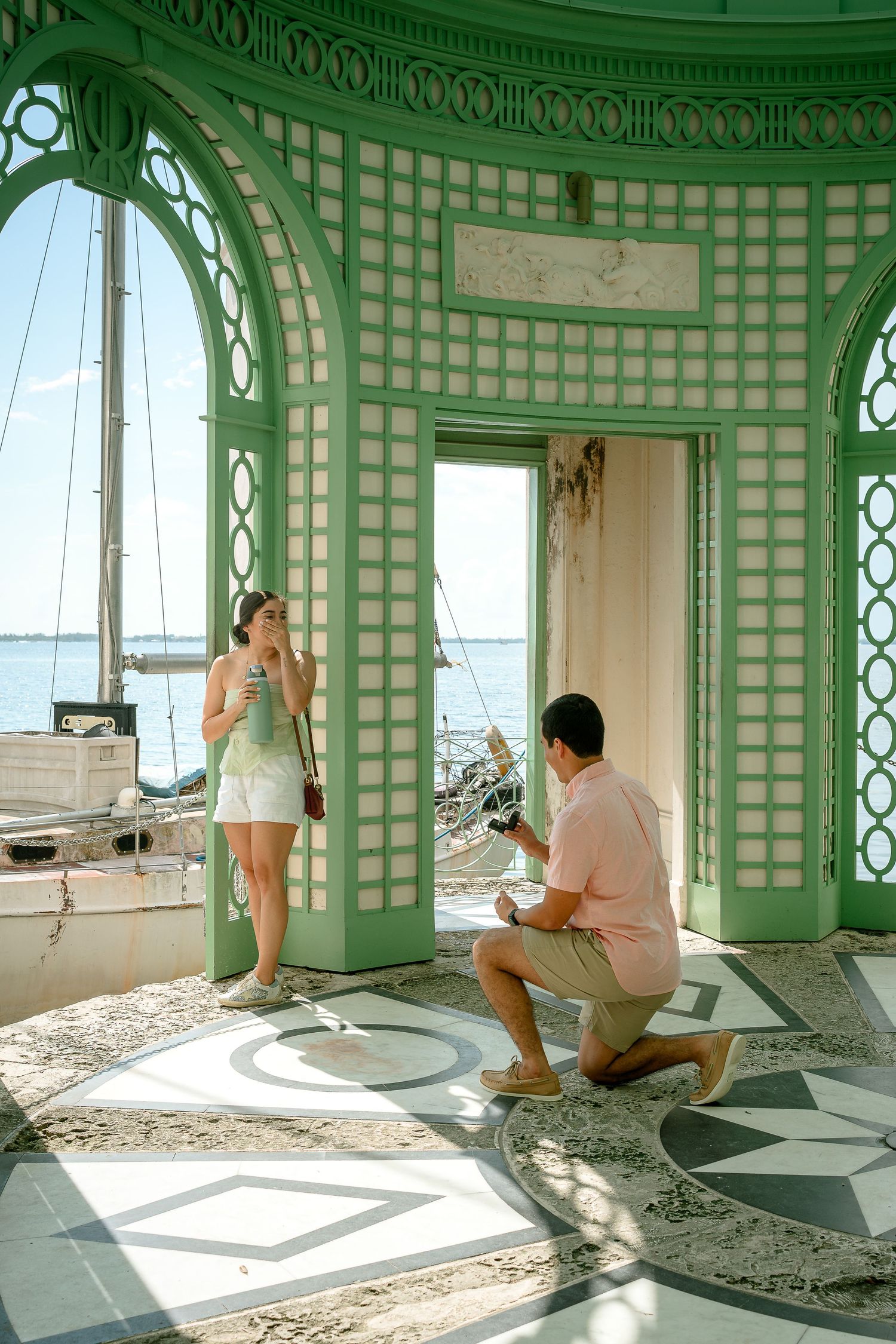 Man proposes on one knee to woman in ornate green pavilion overlooking the ocean with sailboats.