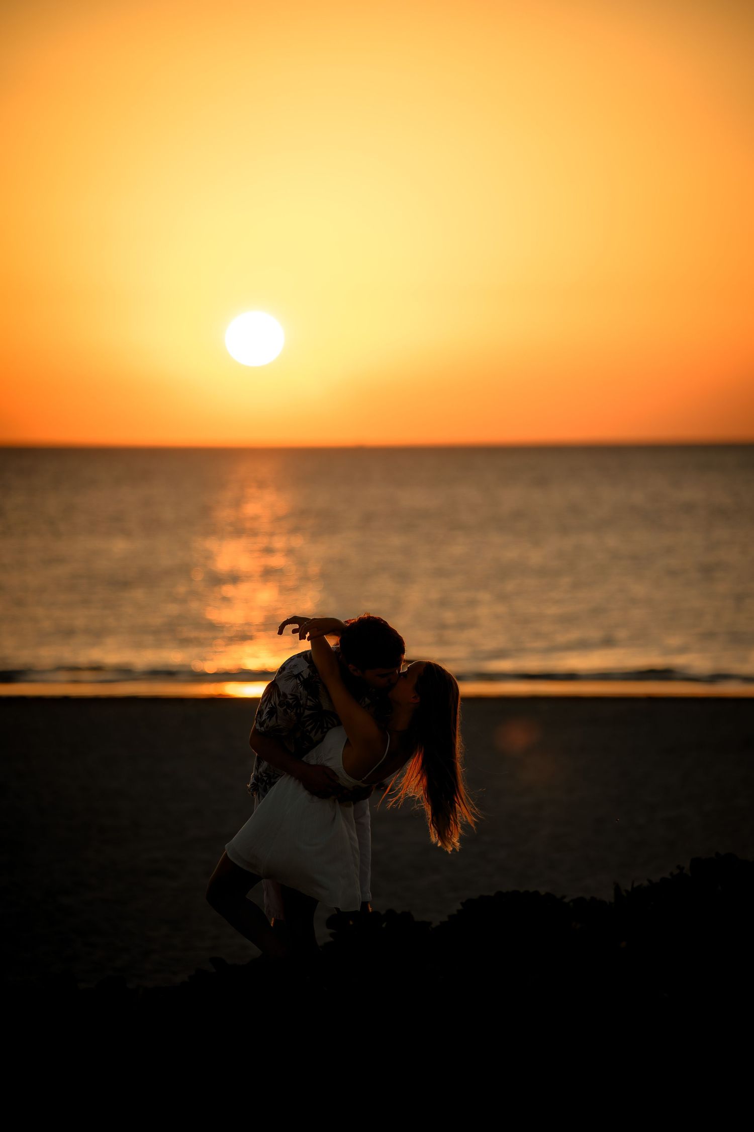 Romantic couple silhouette kissing on beach at golden sunset with sun reflecting on ocean water.