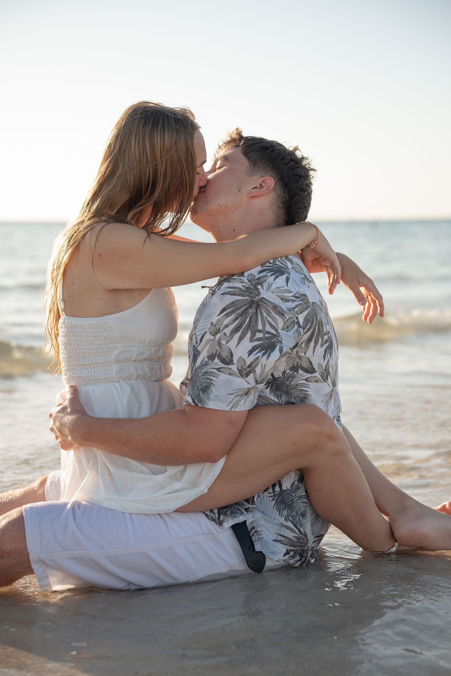Romantic couple kissing while sitting together on a sunny beach, waves gently lapping around them.
