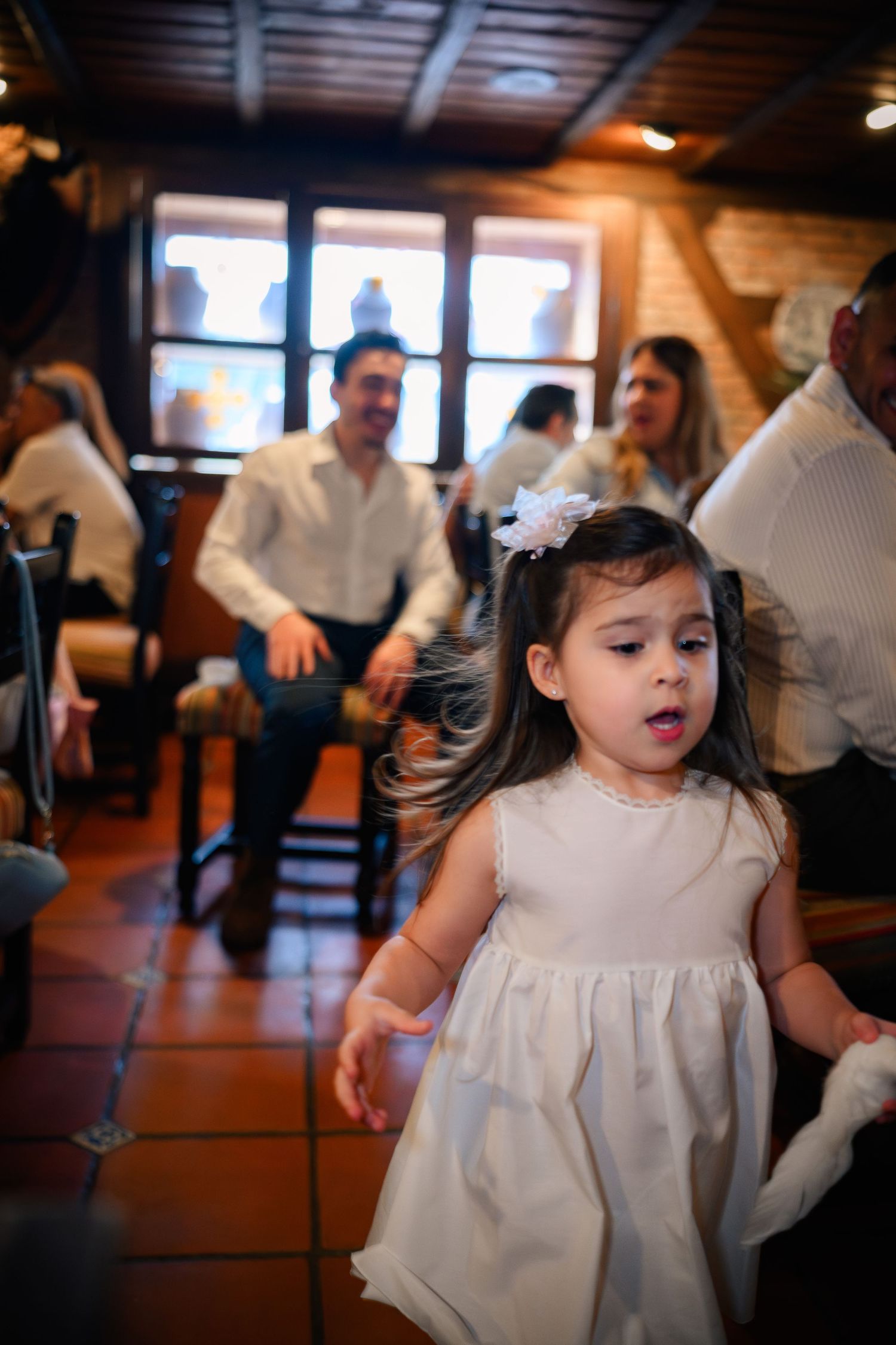 A young child in a white dress dances and spins during a celebration in a rustic indoor venue.