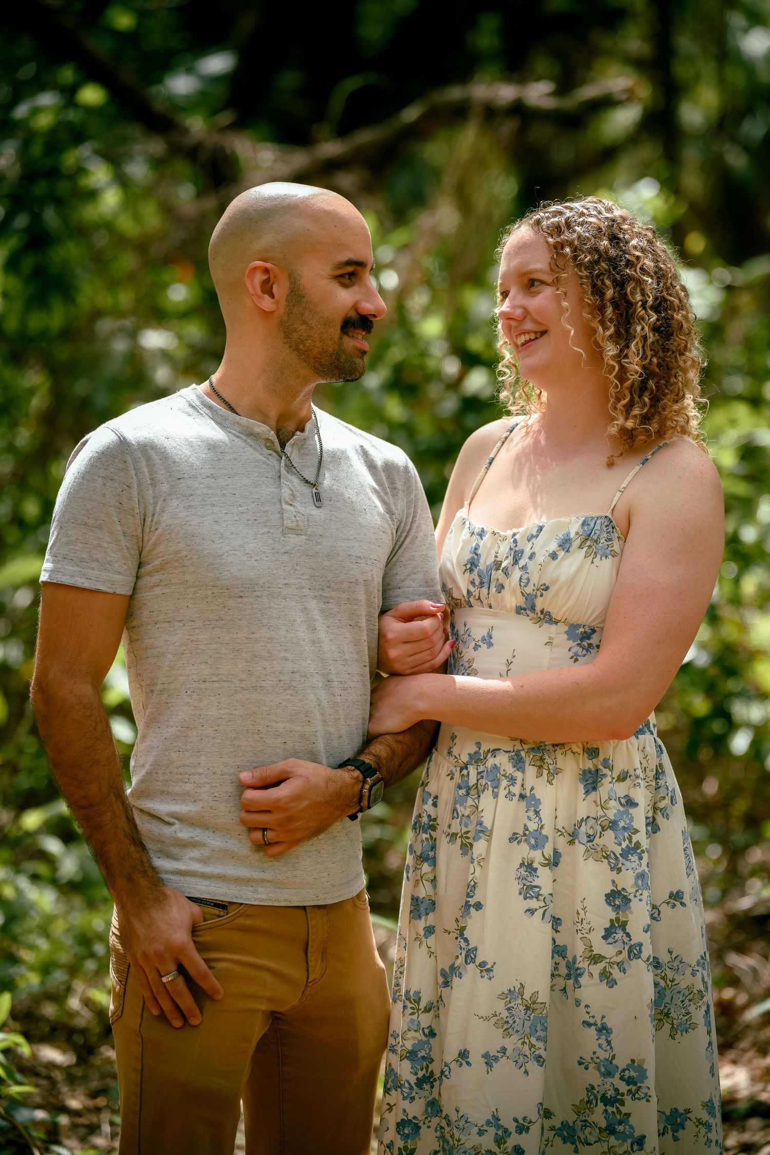 A bald man and curly-haired woman in floral dress smile at each other outdoors in a lush green setting.