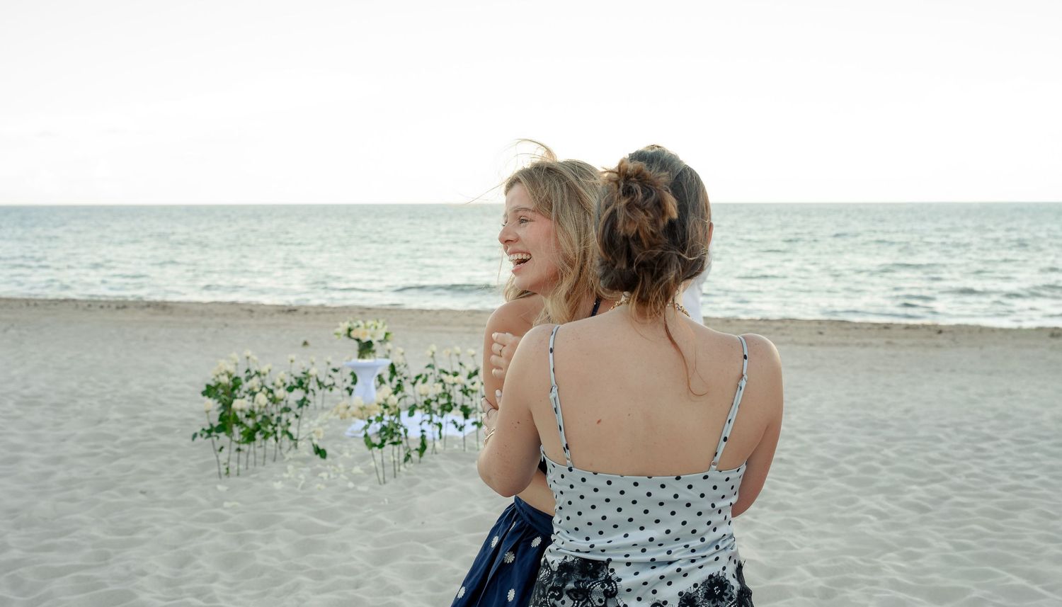Two women in floral dresses embrace on a sandy beach, one holding white flowers, with calm ocean waves in the background.