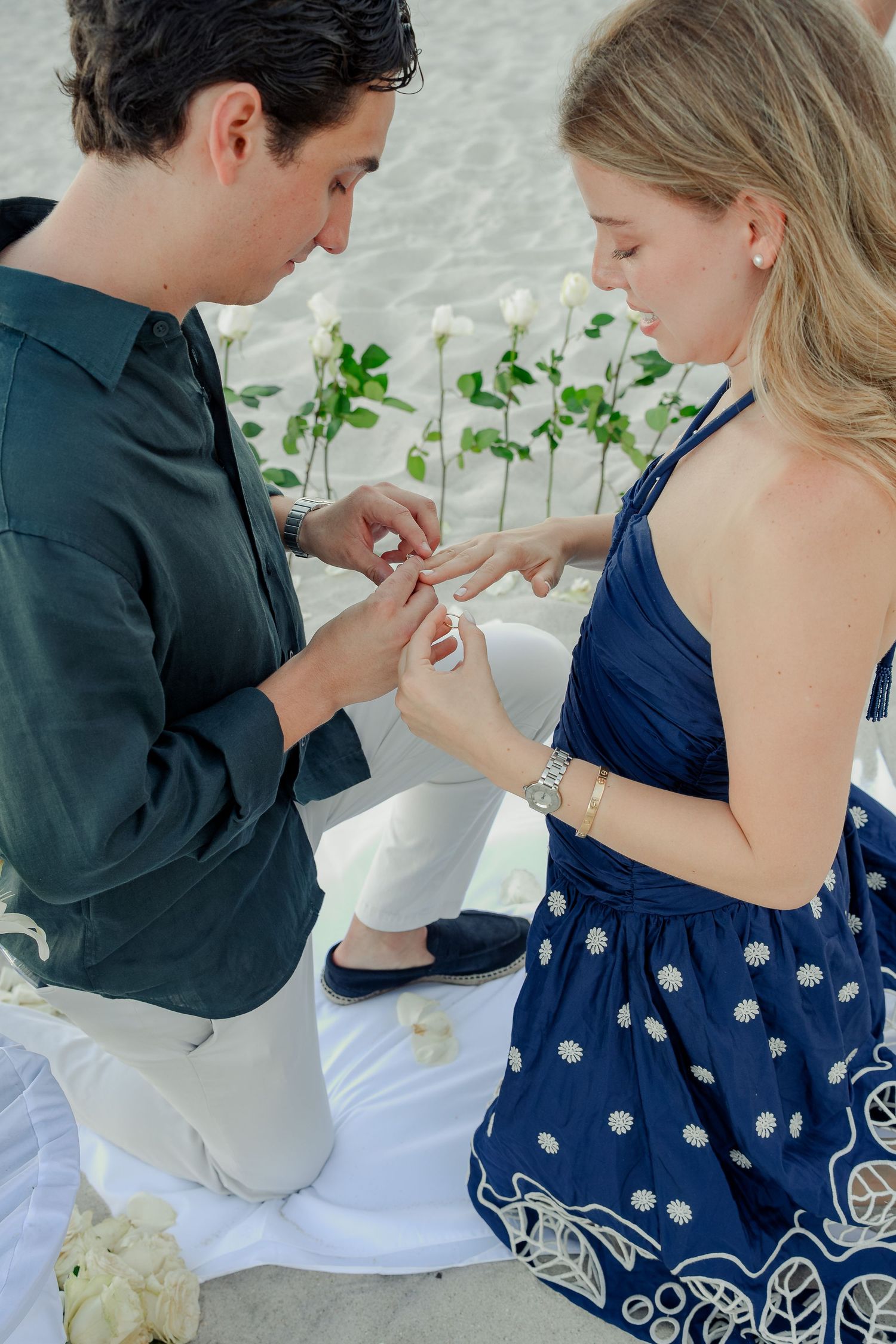 Man kneeling on beach proposing to woman in blue dress, placing engagement ring on her finger.