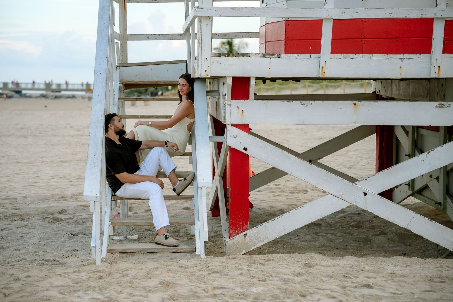 Two people relaxing under a red and white lifeguard stand on a sandy beach at dusk.