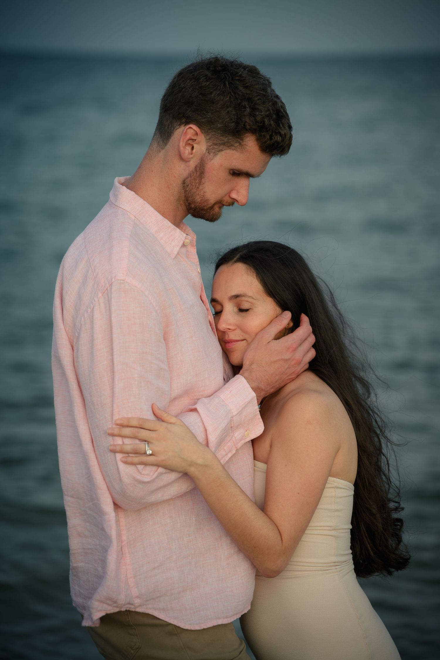 A couple embraces tenderly by the ocean at dusk, woman in strapless dress with engagement ring, man in pink linen shirt.