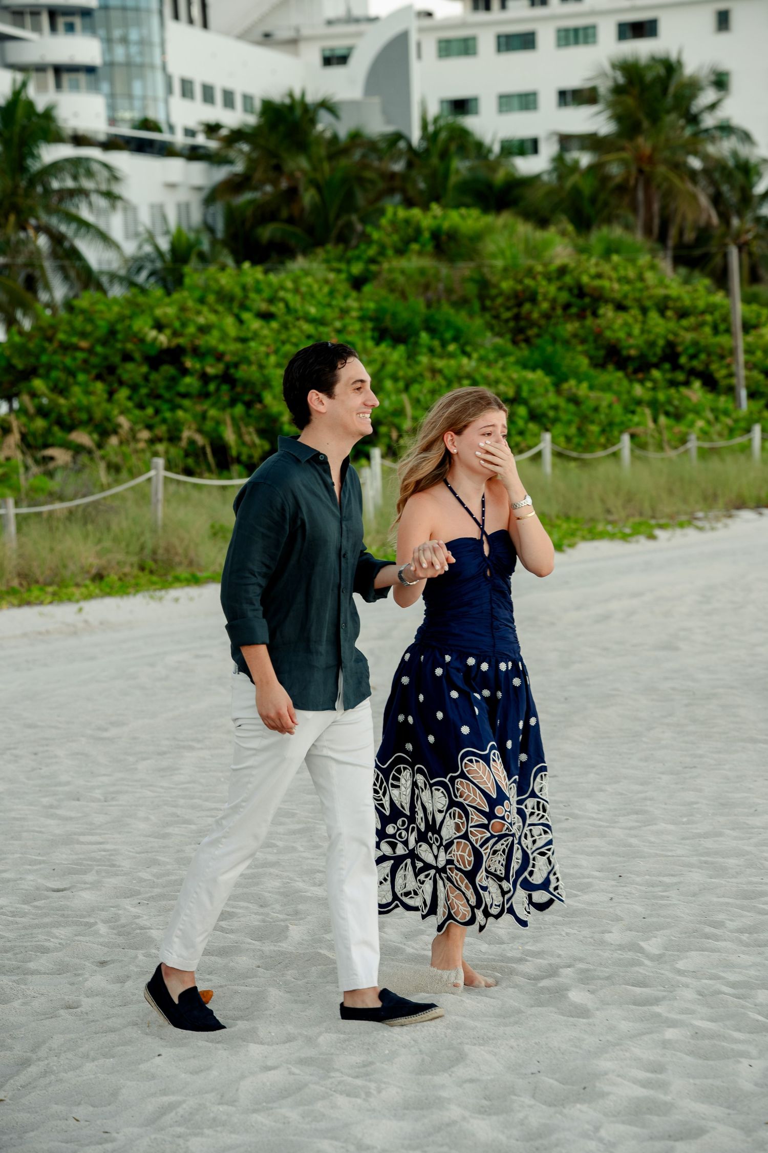 Couple walking on sandy beach, woman in navy floral dress covering mouth, man in dark shirt and white pants, tropical resort background.