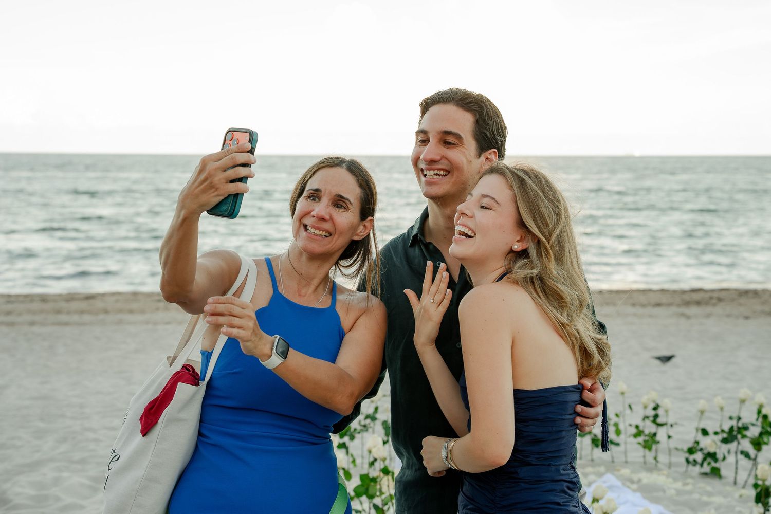Three people taking a selfie on a beach, two women in blue dresses and a man smiling by the ocean.