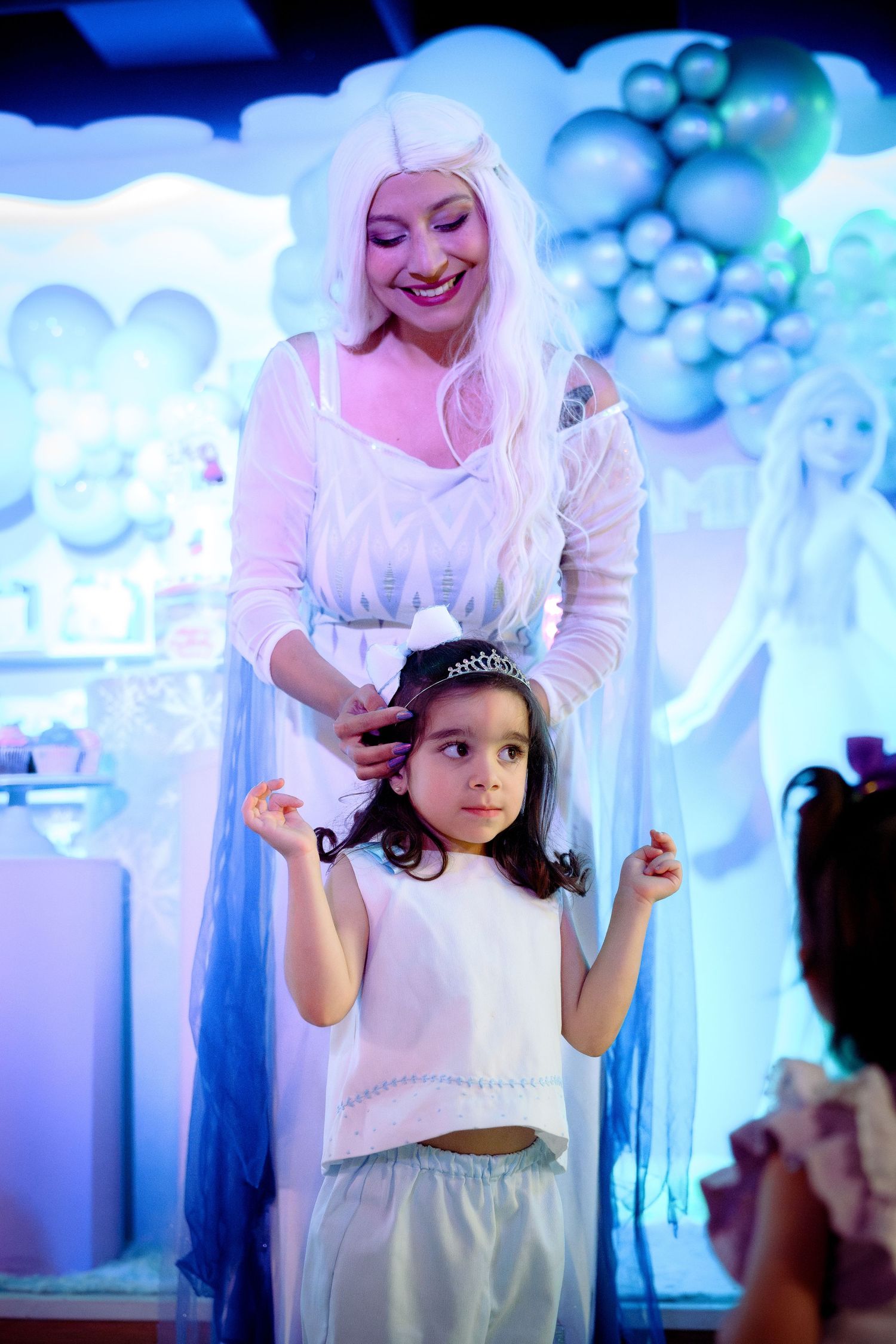 A woman dressed as Elsa from Frozen places a tiara on a little girl at a themed birthday party.