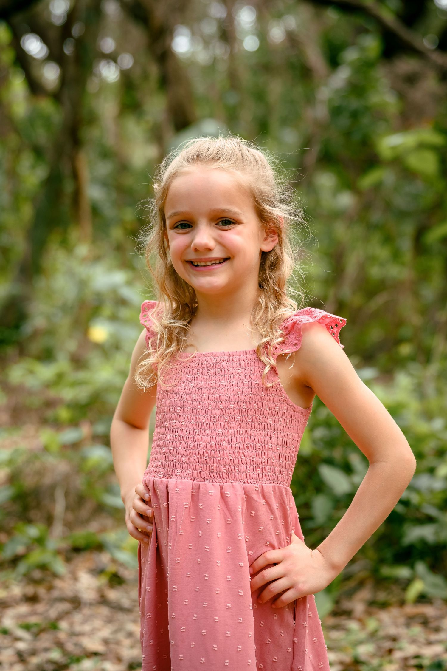 Smiling young girl with blonde hair wearing a pink smocked dress, posing outdoors in a lush green forest setting.