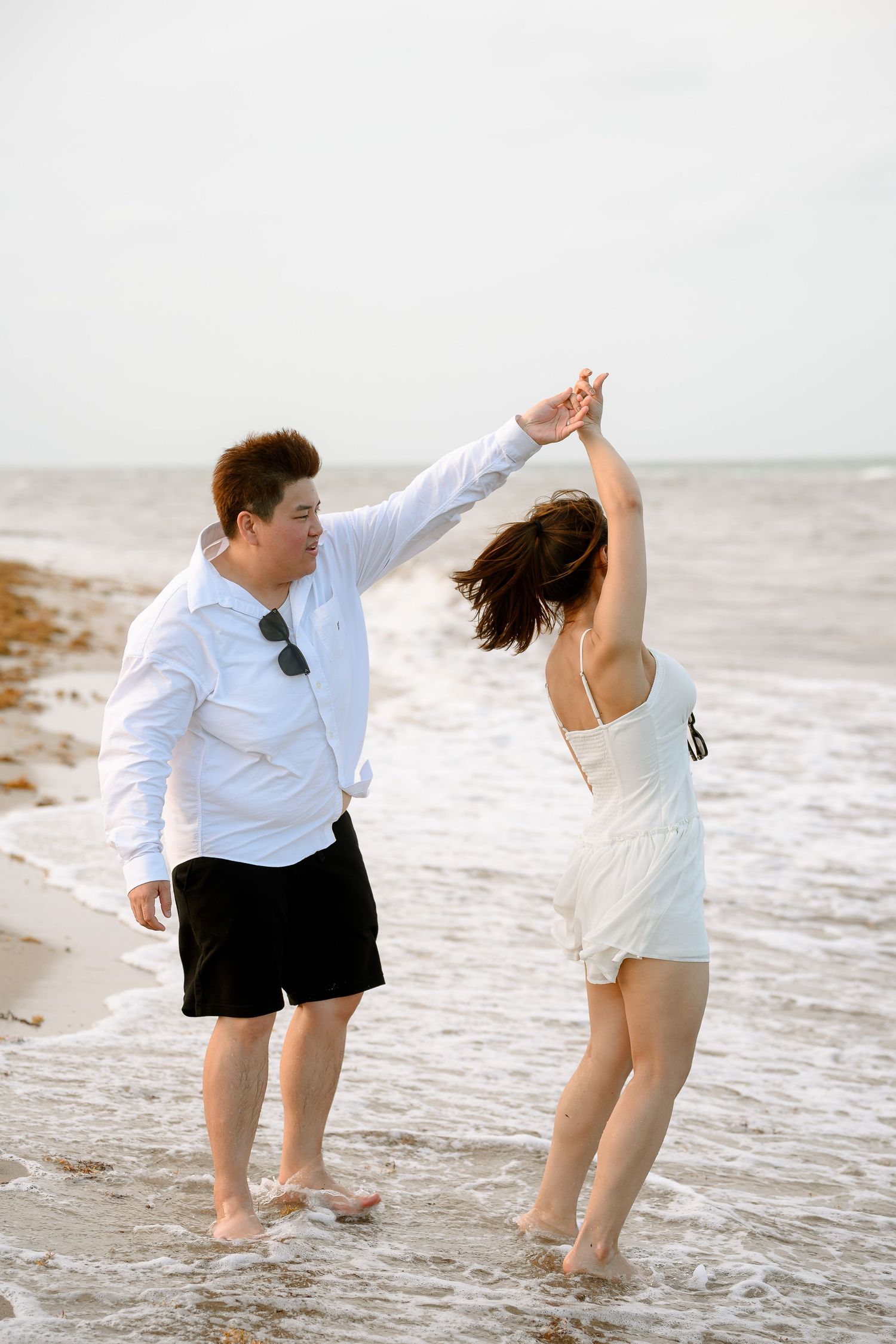 Couple dancing barefoot on beach shoreline, man in white shirt and black shorts spinning woman in white dress.