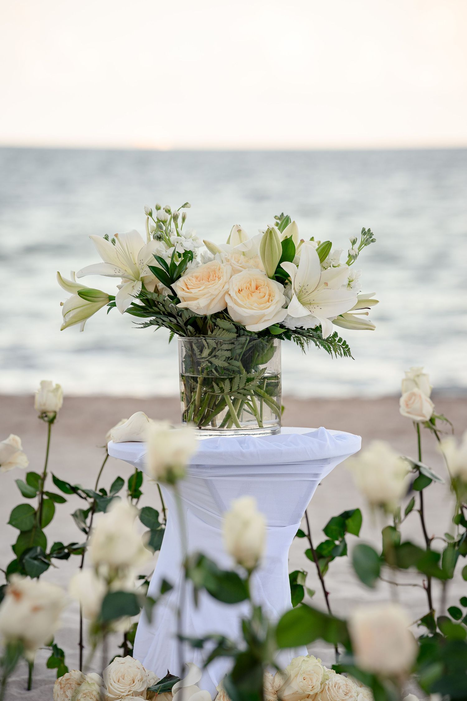 White roses and lilies bouquet in glass vase on draped table at beach wedding ceremony at sunset.