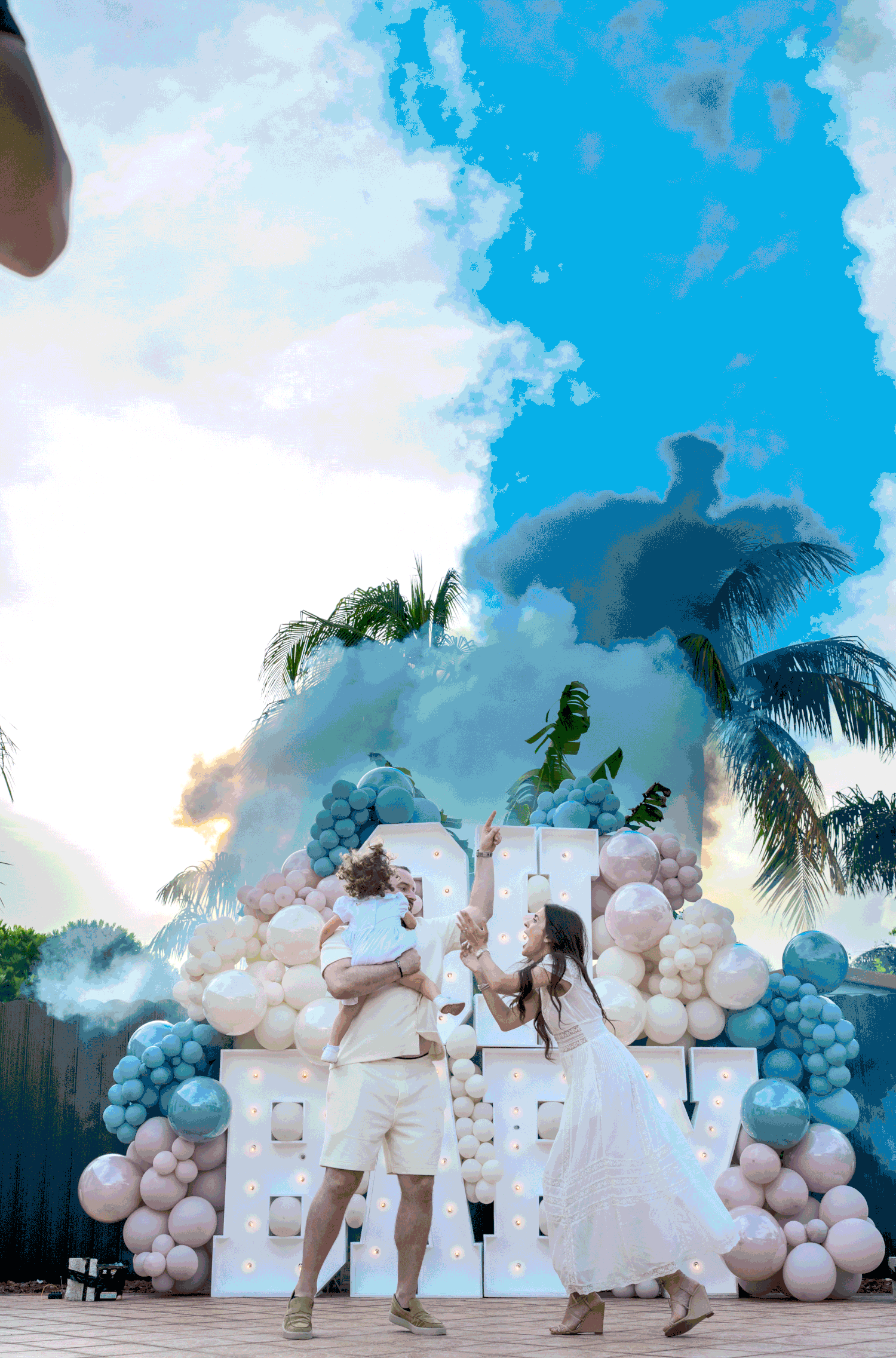 Outdoor gender reveal party setup with white and blue balloon arch and smoke effects against palm trees and blue sky.