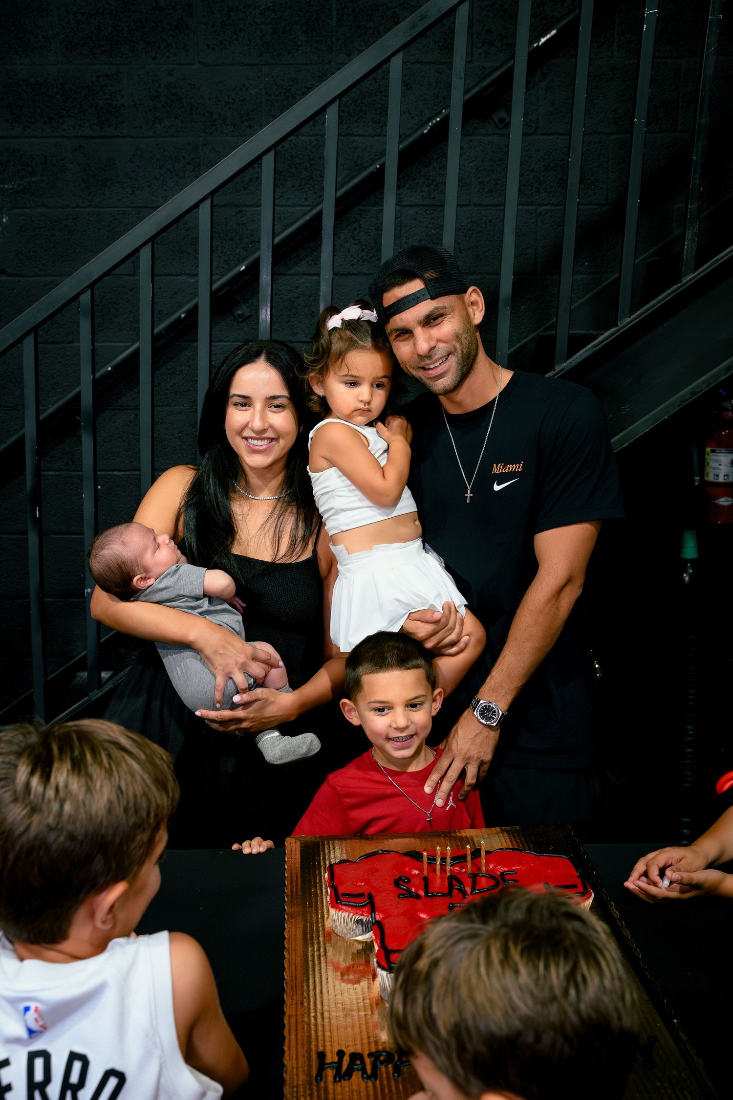 Family posing together at a birthday party with a cake, including parents holding young children near stairs.