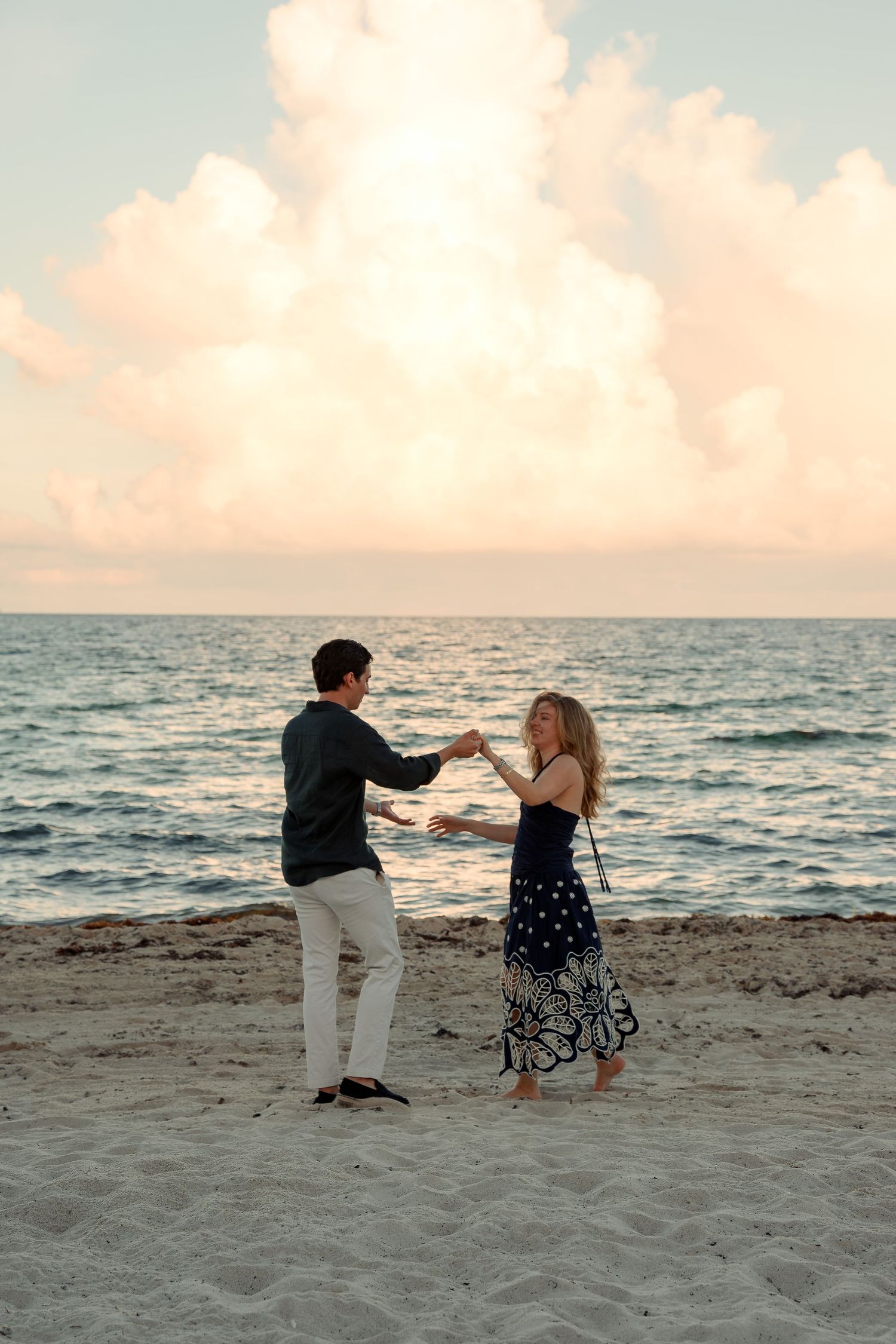 Couple dancing on sandy beach at sunset with pastel clouds and ocean waves in the background.