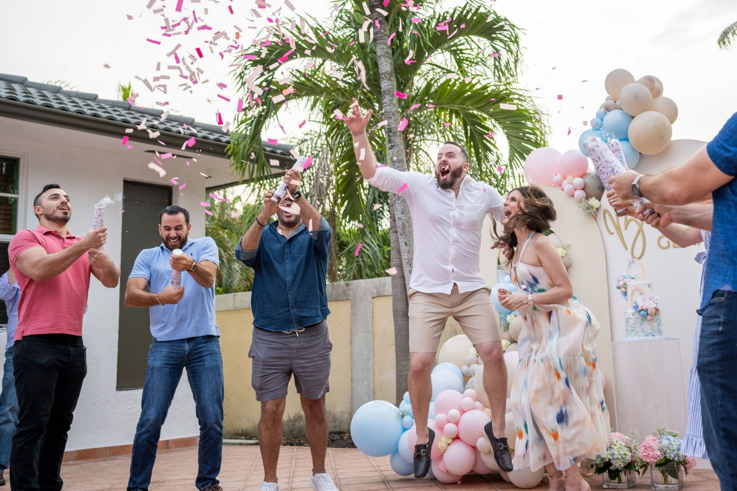 Group of friends celebrating a gender reveal party outdoors with pink confetti, balloons, and joyful expressions.