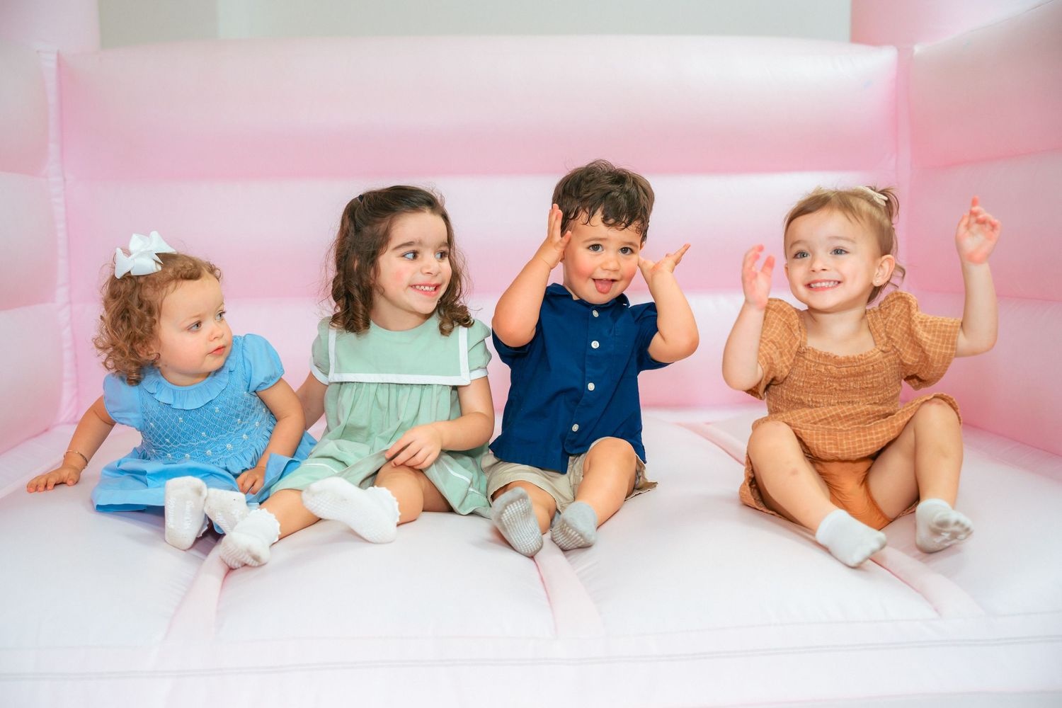 Five children in casual clothing laying and playing together on a white surface against a pink background.