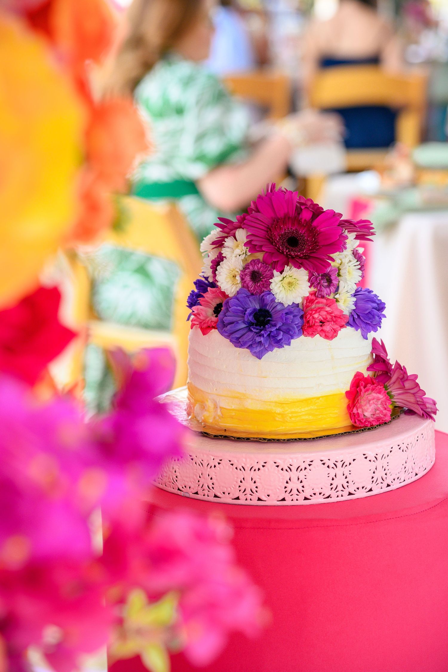 A colorful wedding cake decorated with vibrant purple and pink fresh flowers on a pink tablecloth.