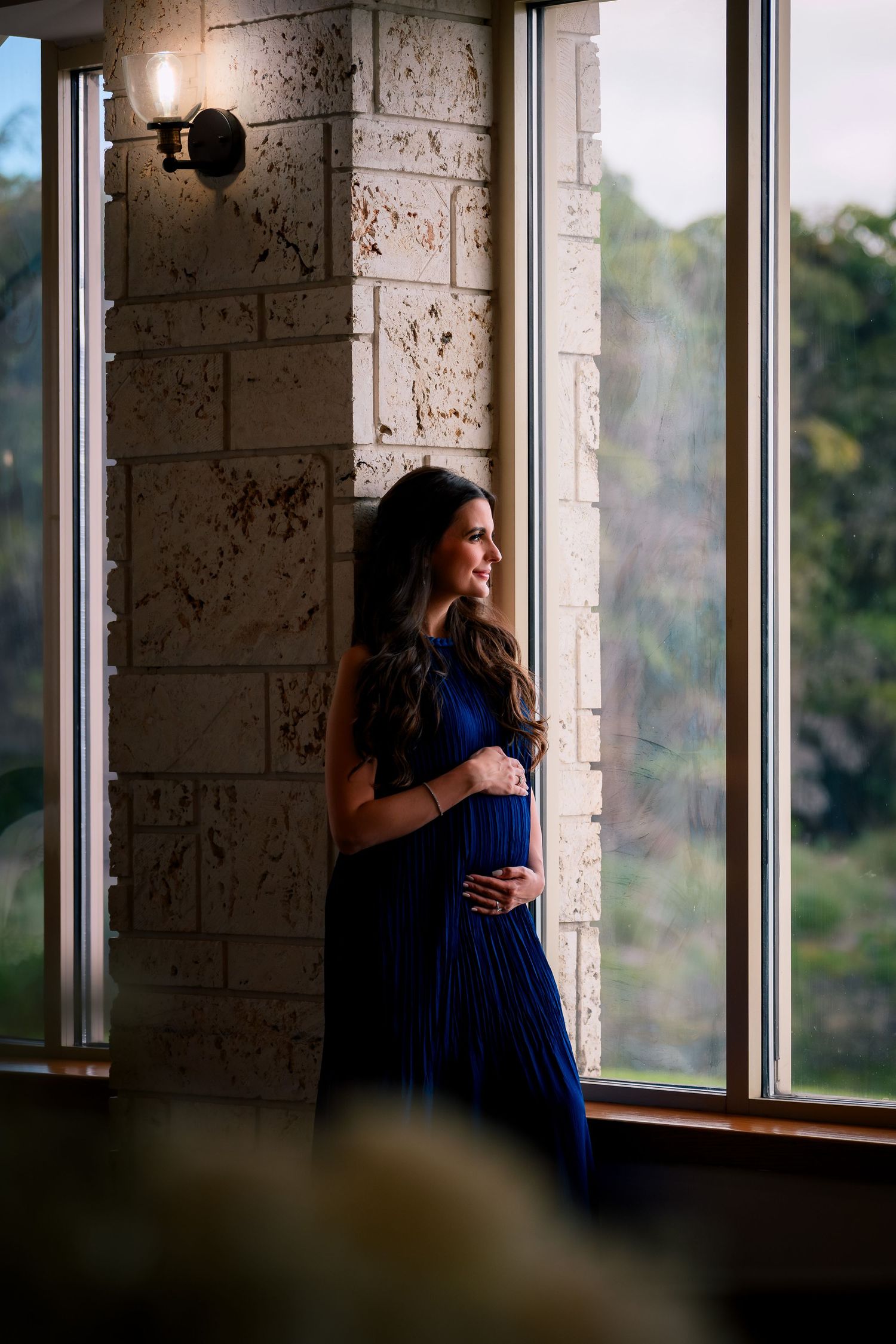 A pregnant woman in a royal blue dress stands by large windows in soft natural light.