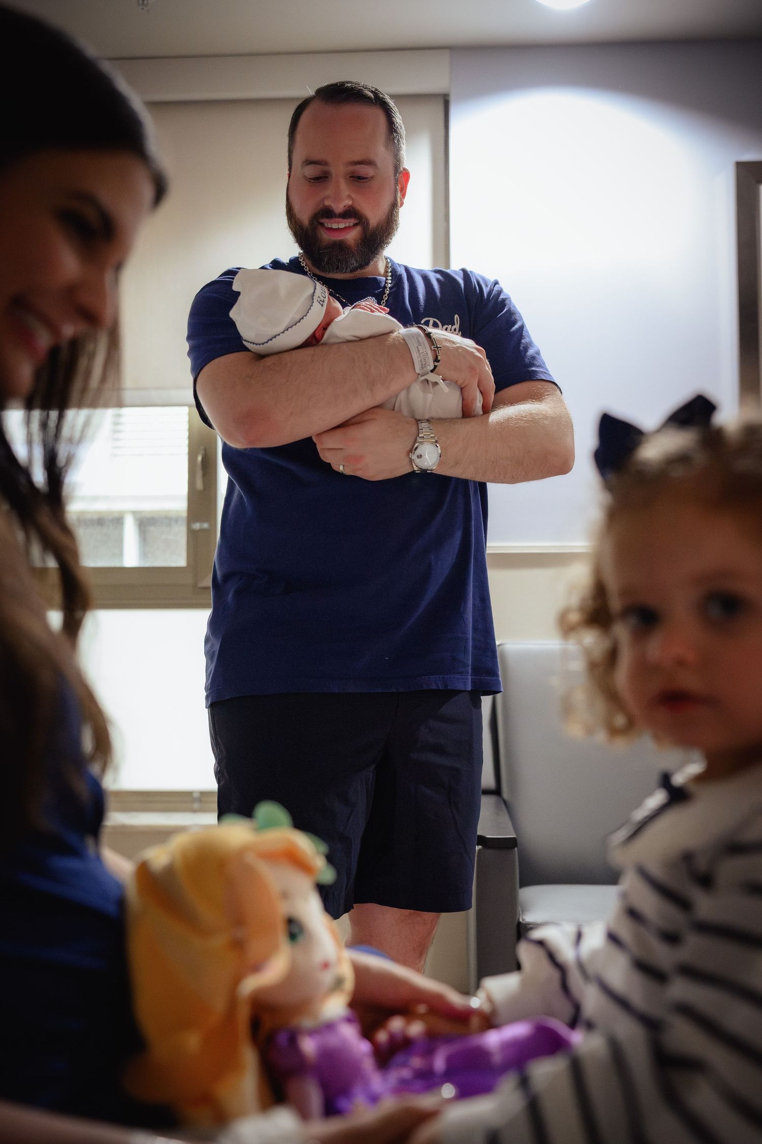 Proud father holding newborn baby in hospital room while toddler daughter plays nearby.