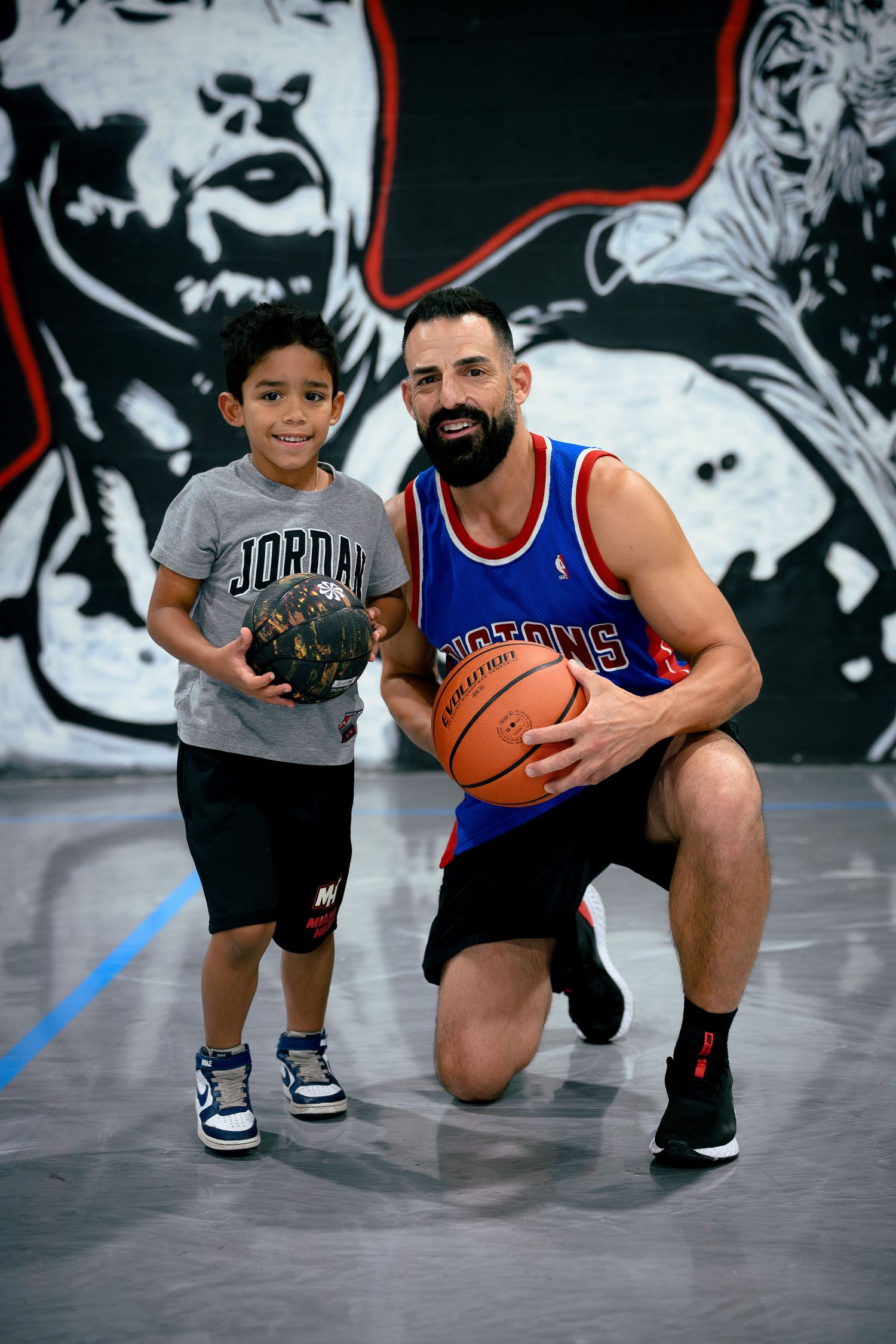 Man in Pistons jersey and boy in Jordan shirt pose with basketballs on court with NBA mural backdrop.