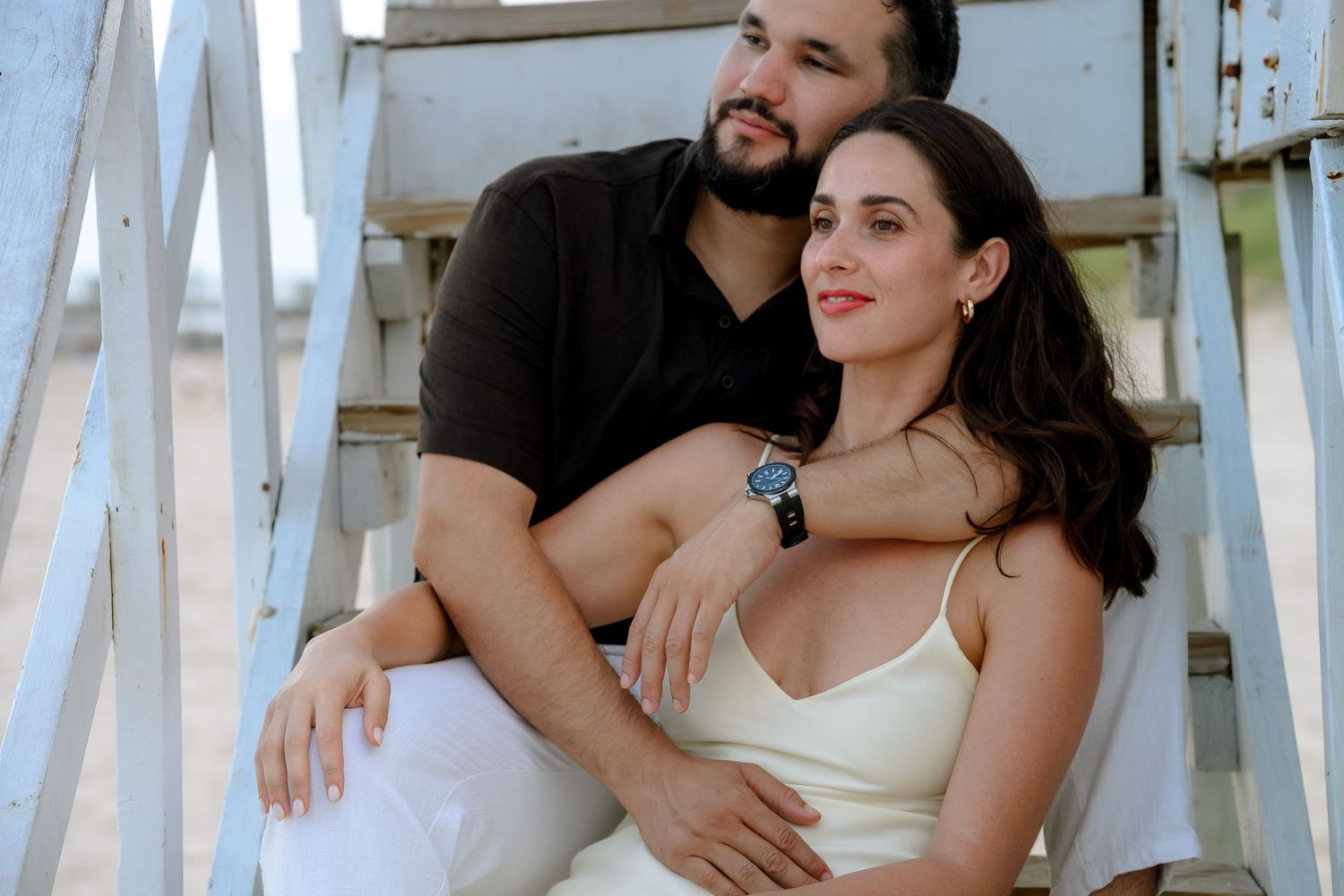 Couple sitting on white beach steps, man in black shirt embracing woman in white dress, romantic outdoor portrait.