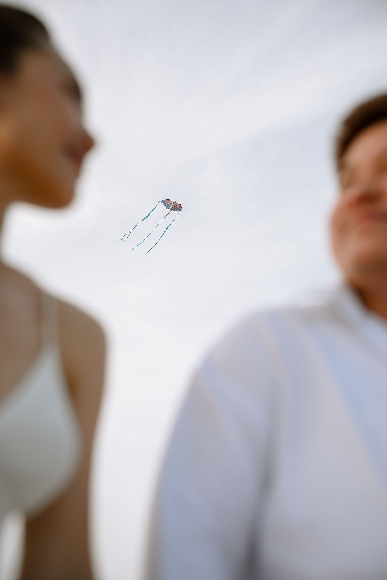 Couple in white standing on beach with colorful kite flying in cloudy sky between them.